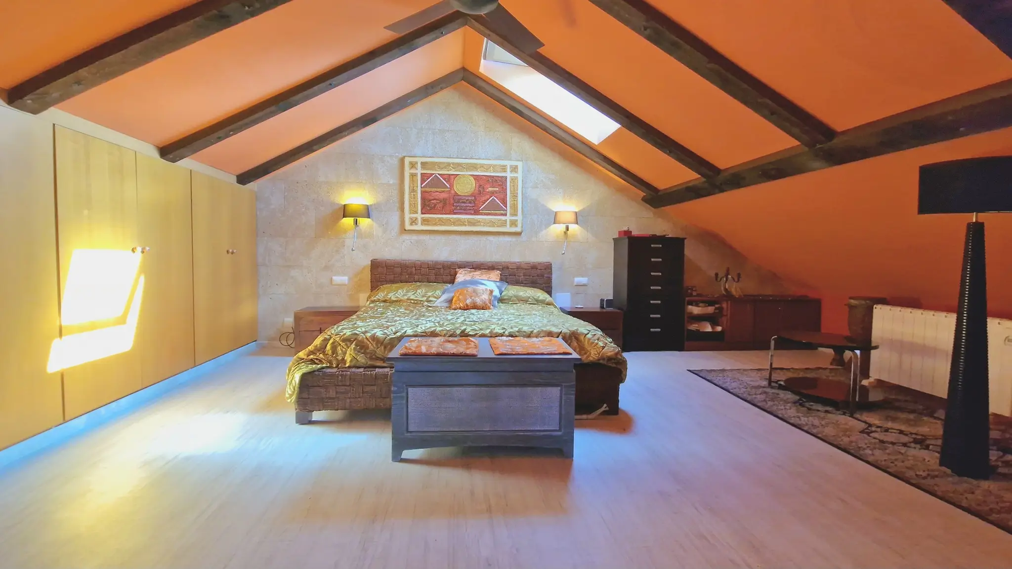Dormitorio abuhardillado con vigas de madera oscura, techos inclinados en tono naranja, pared frontal de piedra y amplios armarios empotrados de madera clara.