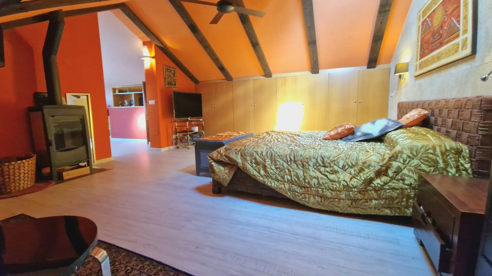 Dormitorio abuhardillado con vigas de madera vista, estufa de leña y paredes terracota. Incluye armarios empotrados, ventilador de techo y cabecero de fibras naturales.