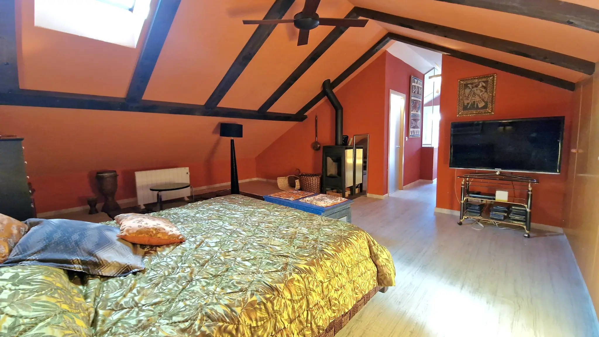 Dormitorio abuhardillado con vigas de madera vista, paredes en tono terracota, estufa de leña y tragaluz que aporta iluminación natural.