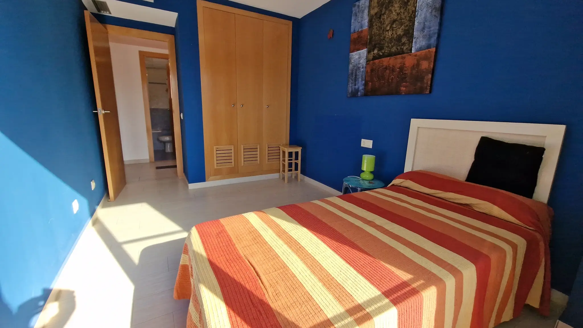 Dormitorio con paredes azul intenso, armario empotrado de madera y suelos cerámicos. La estancia cuenta con cama individual y acceso directo a un baño privado.
