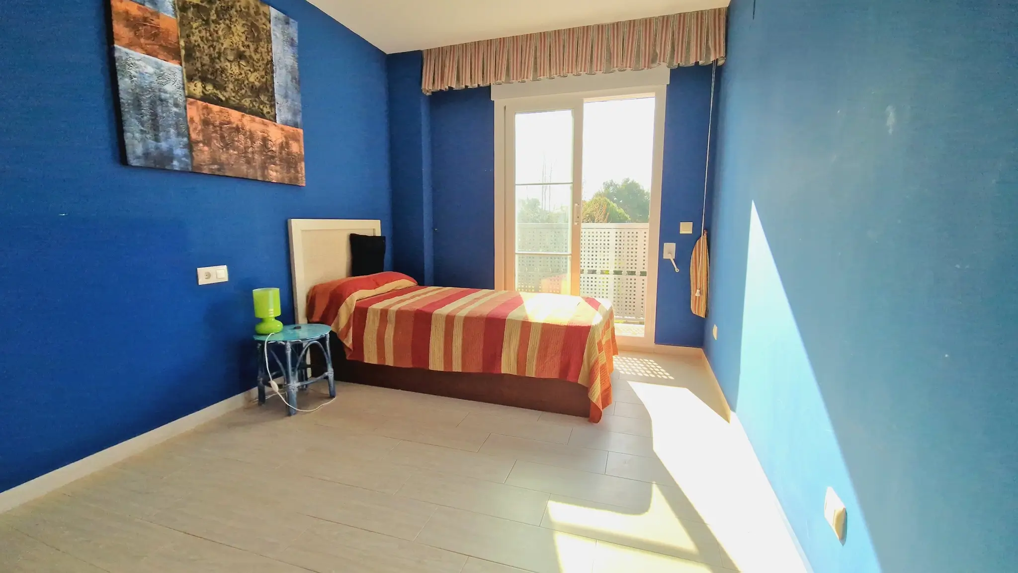 Dormitorio con paredes azul intenso, cama individual con ropa de cama a rayas y acceso directo a terraza mediante puertas correderas.