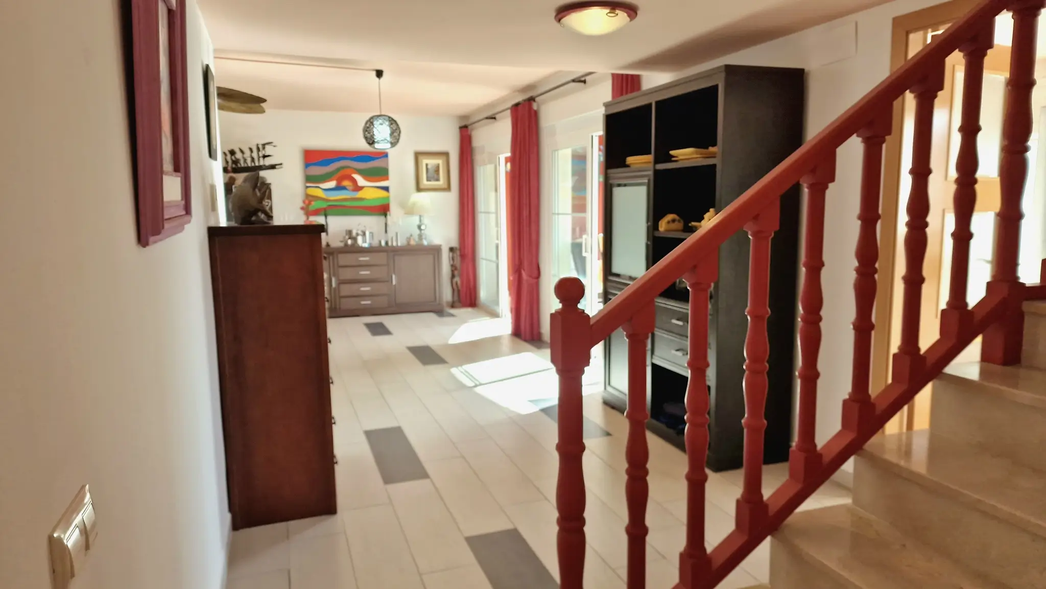 Pasillo con barandilla de madera roja y escalera de mármol, que conecta con un salón luminoso con suelos cerámicos, mobiliario oscuro y cortinas rojas.
