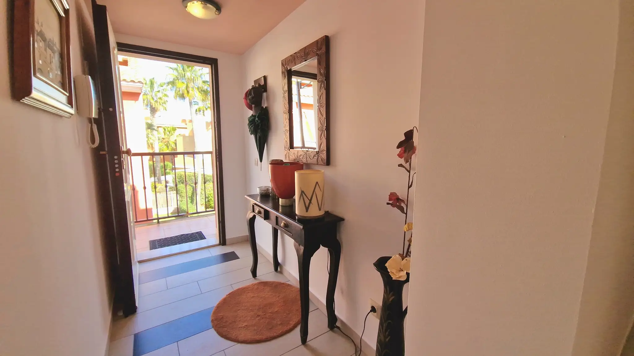 Recibidor con consola de madera oscura, espejo decorativo y suelos cerámicos, con acceso directo a un balcón con vistas a palmeras y luz natural.