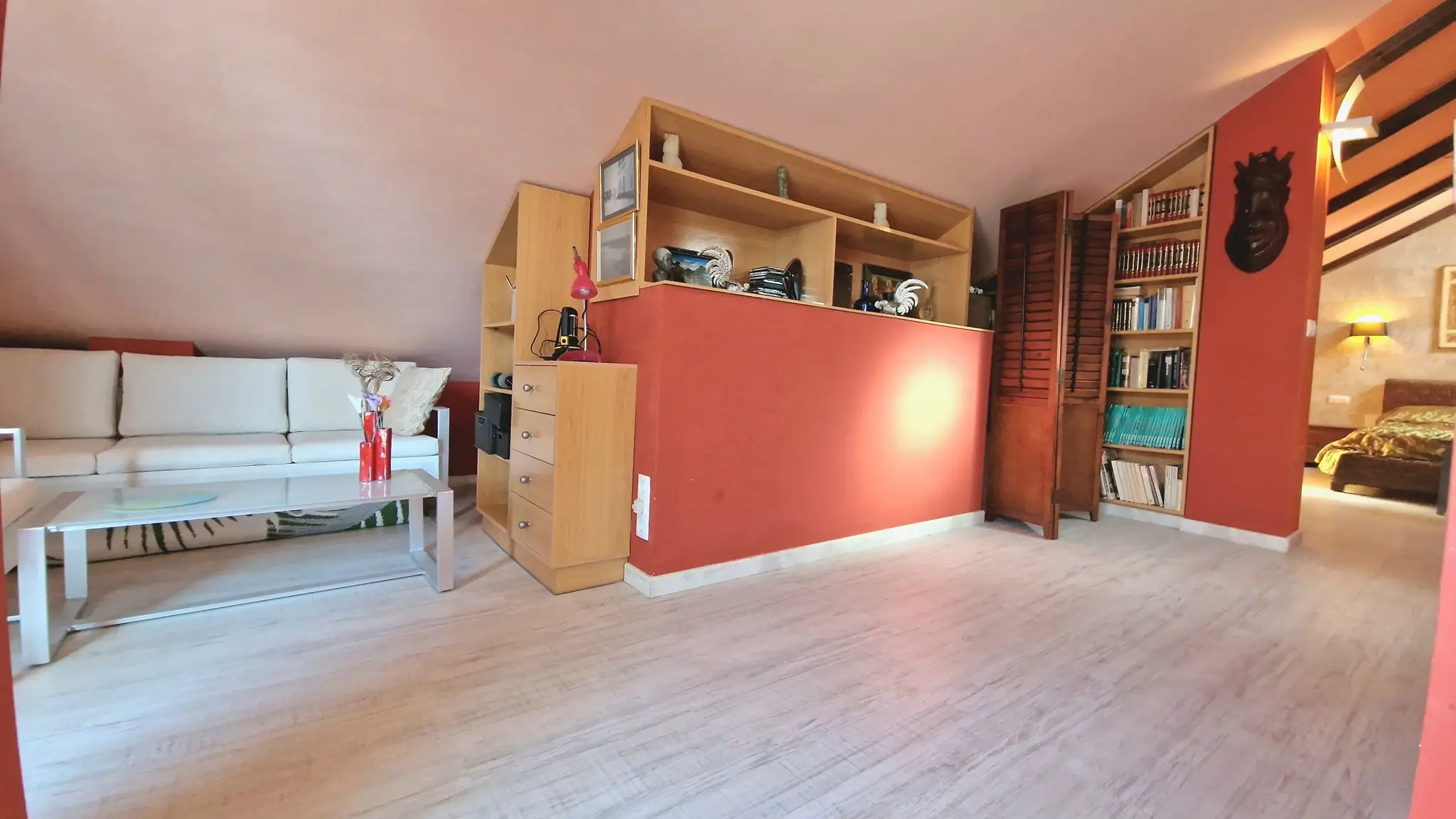 Sala de estar abuhardillada con paredes en tono terracota, estanterías de madera integradas y suelos de laminado claro.