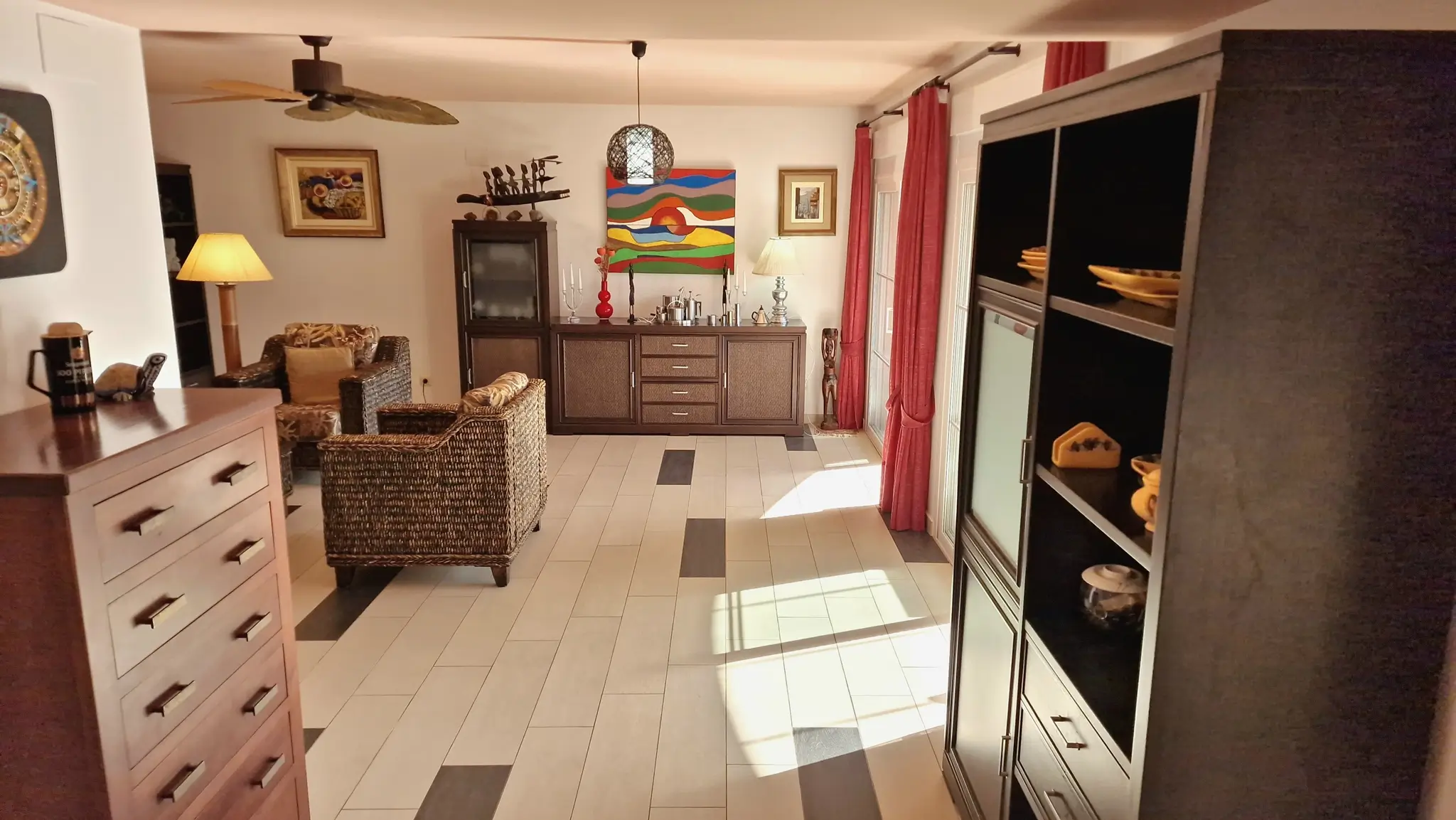 Salón con sillones de mimbre, mobiliario de madera oscura y suelo cerámico con detalles en contraste. La estancia incluye arte colorido y luz natural lateral.