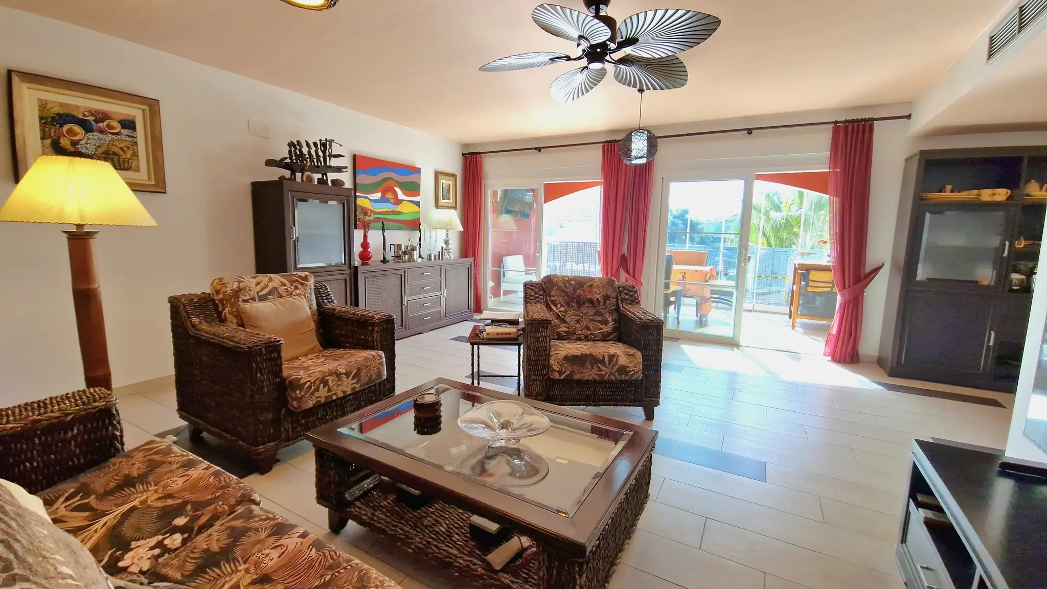 Salón luminoso con muebles de mimbre y motivos tropicales, ventilador de techo estilo palmera y acceso directo a la terraza mediante ventanales correderos.