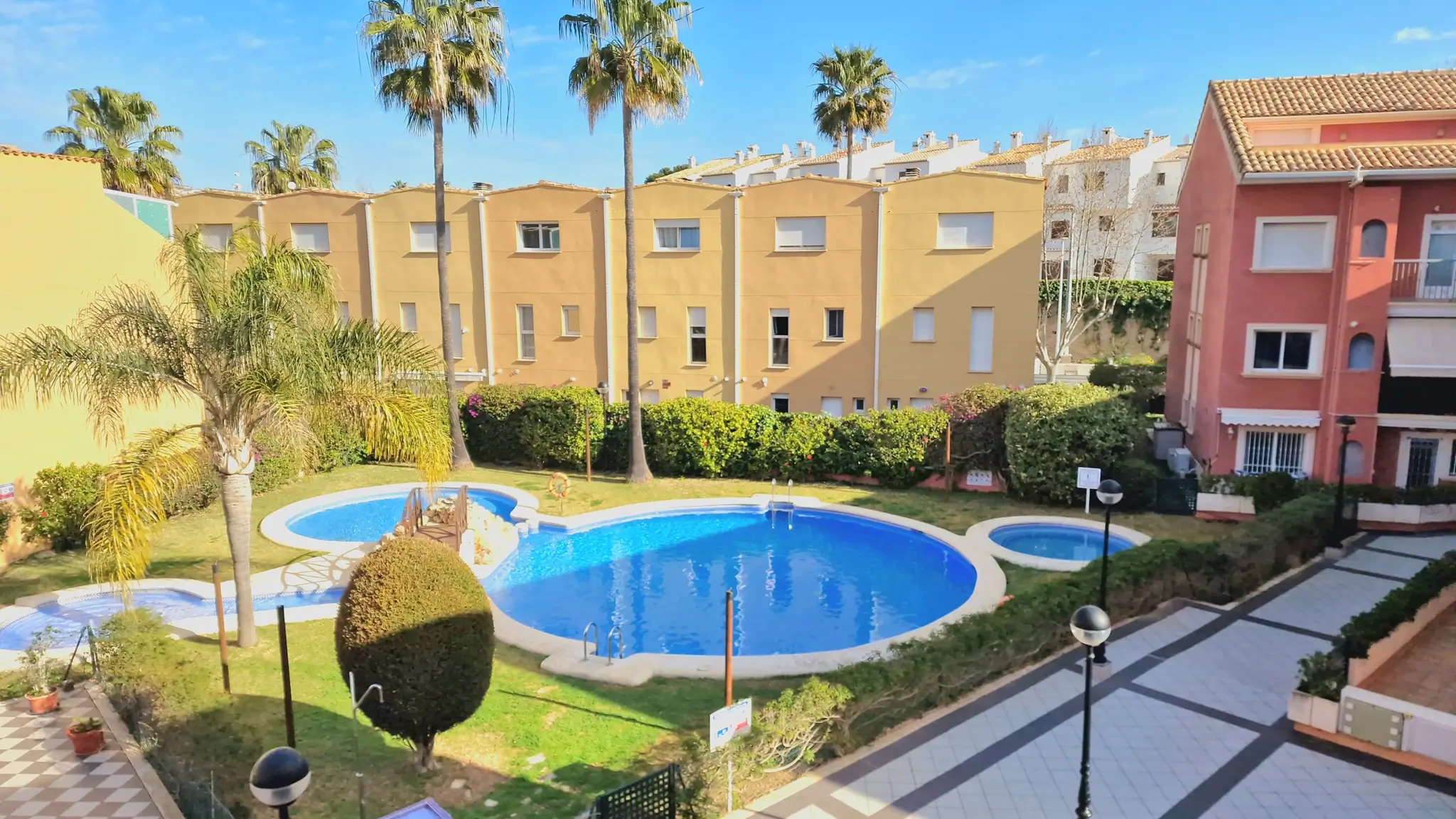 Zona comunitaria con piscinas de diseño curvo, césped cuidado y palmeras, rodeada de edificios residenciales de estilo mediterráneo.
