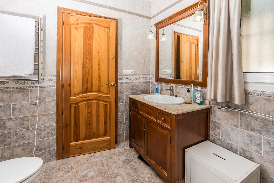 Baño con mueble de lavabo de madera, encimera de piedra y espejo a juego. Incluye puerta de madera natural, toallero radiador y paredes revestidas con azulejos decorativos.