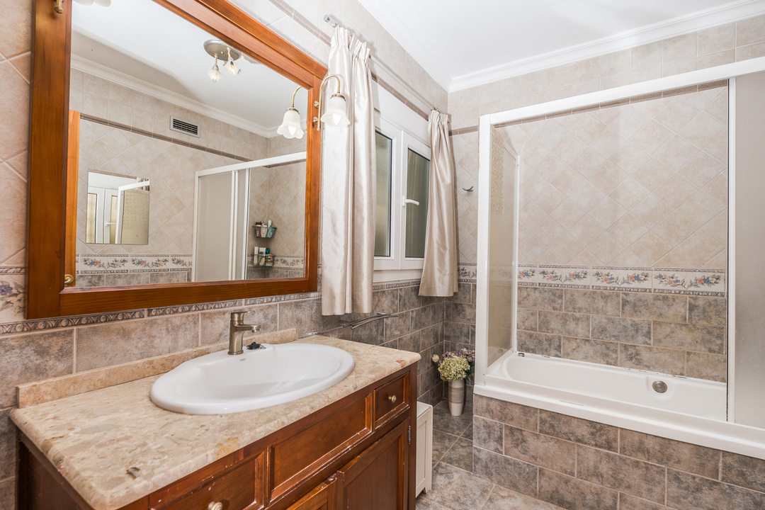 Baño con mueble de madera y encimera de mármol, espejo de gran formato con marco de madera y bañera integrada con mampara corredera y revestimiento cerámico.