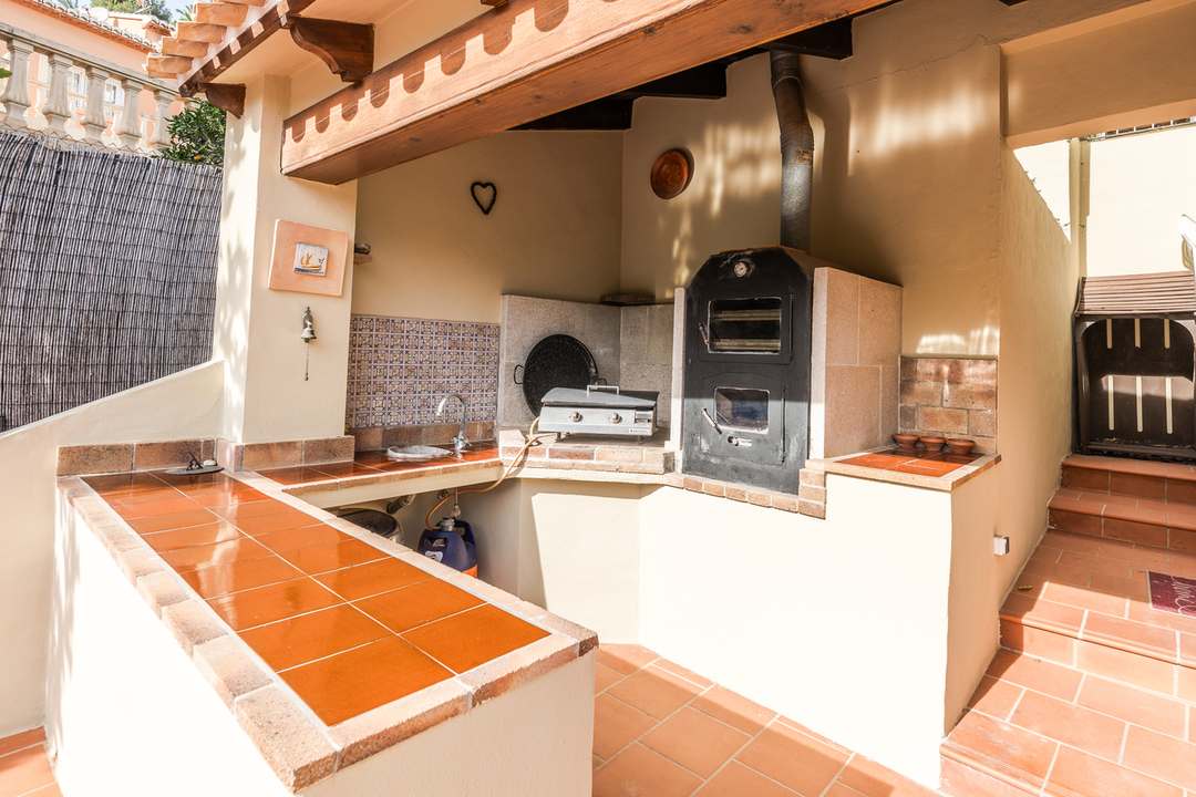 Cocina exterior con horno de leña, encimeras de baldosas de terracota y salpicadero decorativo bajo un porche de vigas de madera.