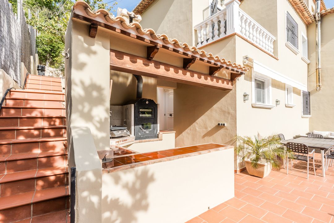 Cocina exterior con horno de leña integrado, encimeras de azulejo terracota y vigas de madera, situada junto a la terraza comedor y escaleras laterales.