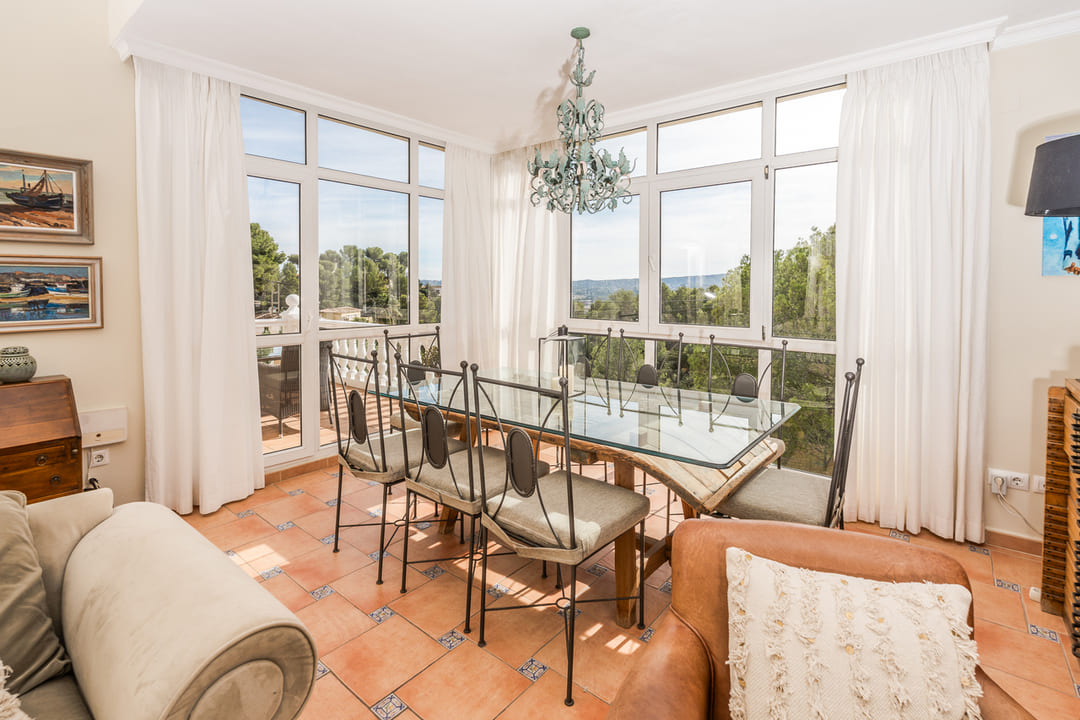 Comedor luminoso con mesa de cristal, sillas de hierro forjado y suelo de baldosas cerámicas. Amplios ventanales ofrecen vistas despejadas al entorno arbolado.
