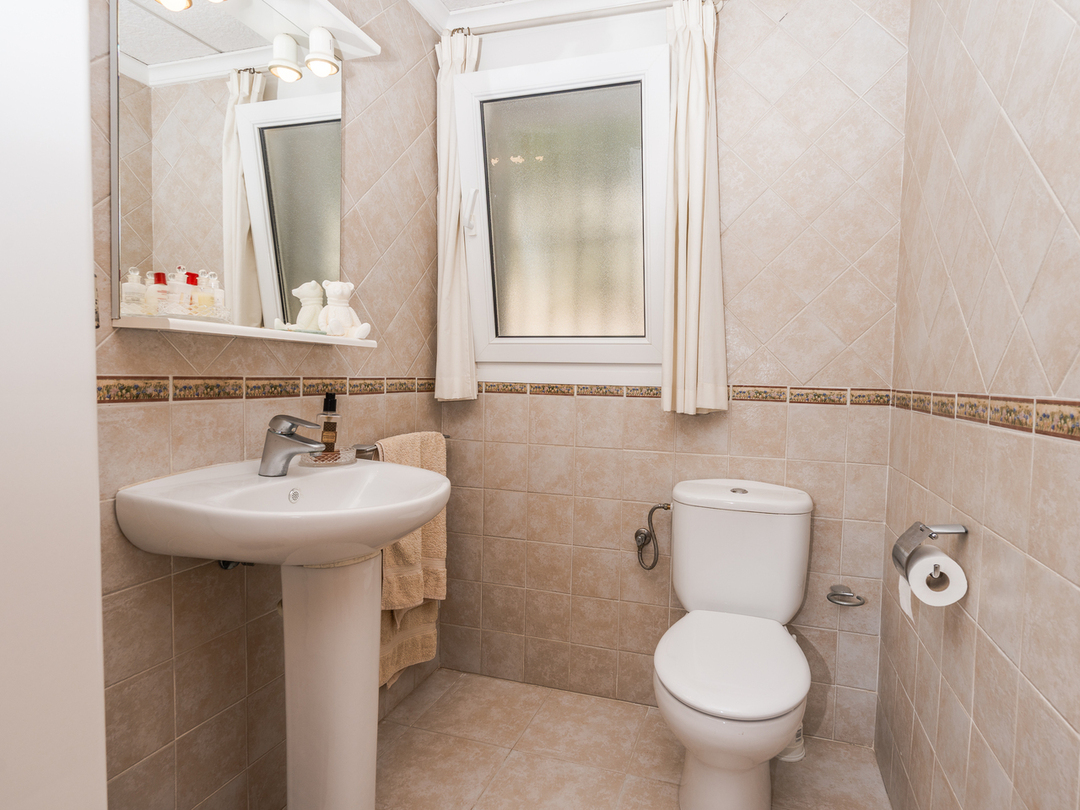 Cuarto de baño con lavabo de pedestal, inodoro de cerámica blanca y paredes alicatadas en tonos beige con cenefa decorativa y ventana.