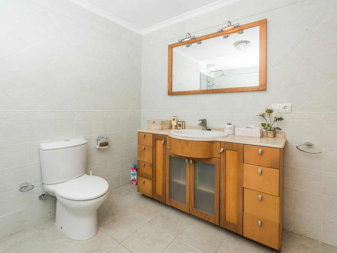 Cuarto de baño con mueble de madera de varios cajones, lavabo integrado, espejo con marco de madera e iluminación superior y paredes alicatadas en tonos claros.