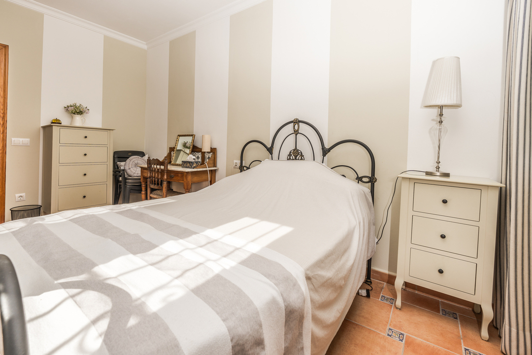 Dormitorio con cabecero de forja, papel pintado a rayas y suelo de terracota. Incluye una mesita de noche crema y un tocador de madera clásica.