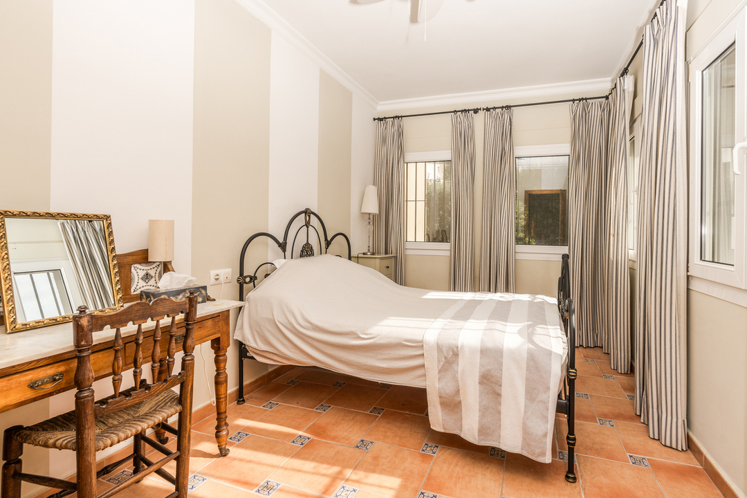 Dormitorio con cama de forja negra, paredes con papel pintado a rayas y suelos de terracota con azulejos decorativos. Incluye un escritorio de madera clásica con espejo.