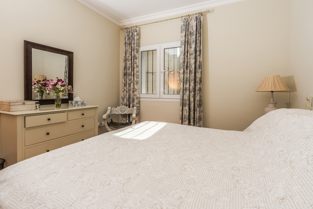Dormitorio con cama doble, cómoda de madera con espejo, cortinas con estampado floral y ventana con rejas de seguridad.