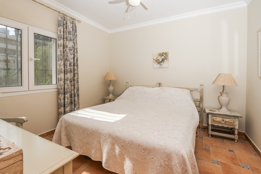 Dormitorio con suelo de terracota y detalles azules, mesitas de noche de madera decapada y luz natural mediante ventana con cortinas de estilo toile.