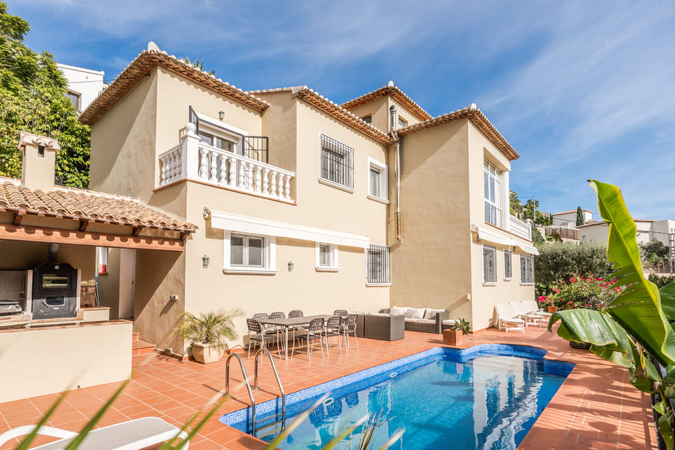 Fachada de villa mediterránea con piscina privada, terraza de terracota, comedor exterior y cocina de verano integrada bajo porche de madera.