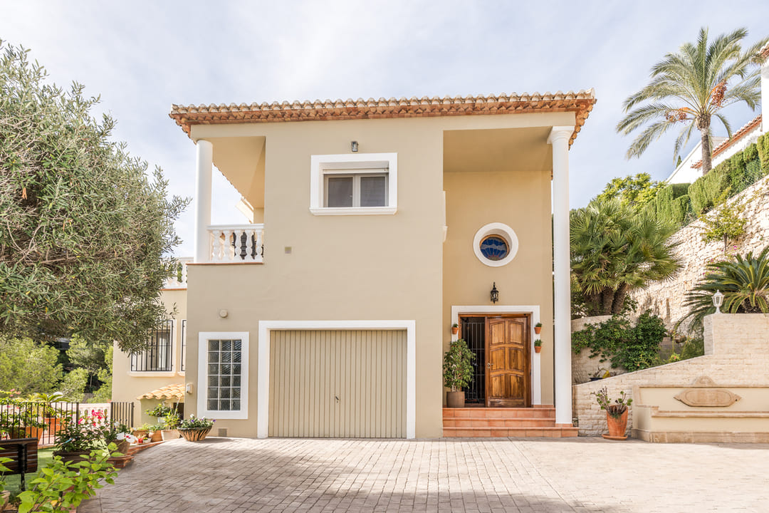 Fachada exterior de una villa de dos niveles con acabado en tonos arena, garaje integrado, puerta de madera y entrada pavimentada rodeada de vegetación mediterránea.