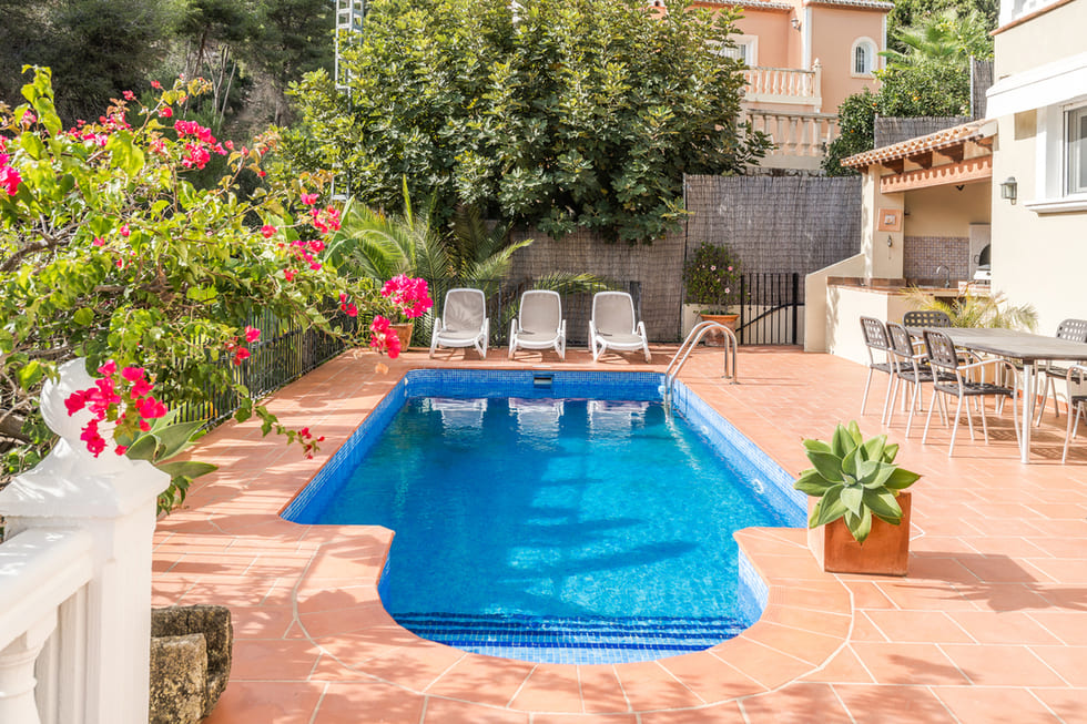 Piscina rectangular de gresite azul rodeada por una terraza de terracota, con zona de hamacas, comedor exterior y vegetación mediterránea con buganvillas.