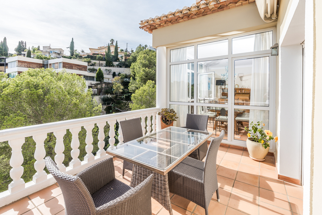 Terraza con suelo de terracota, balaustrada blanca y comedor exterior de cristal. Ofrece vistas a la ladera arbolada y a las villas residenciales circundantes.