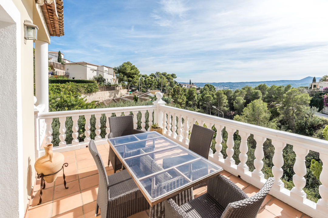 Terraza elevada con mesa de cristal, sillas de mimbre y balaustrada blanca, ofreciendo vistas despejadas al valle de pinos y las colinas circundantes.