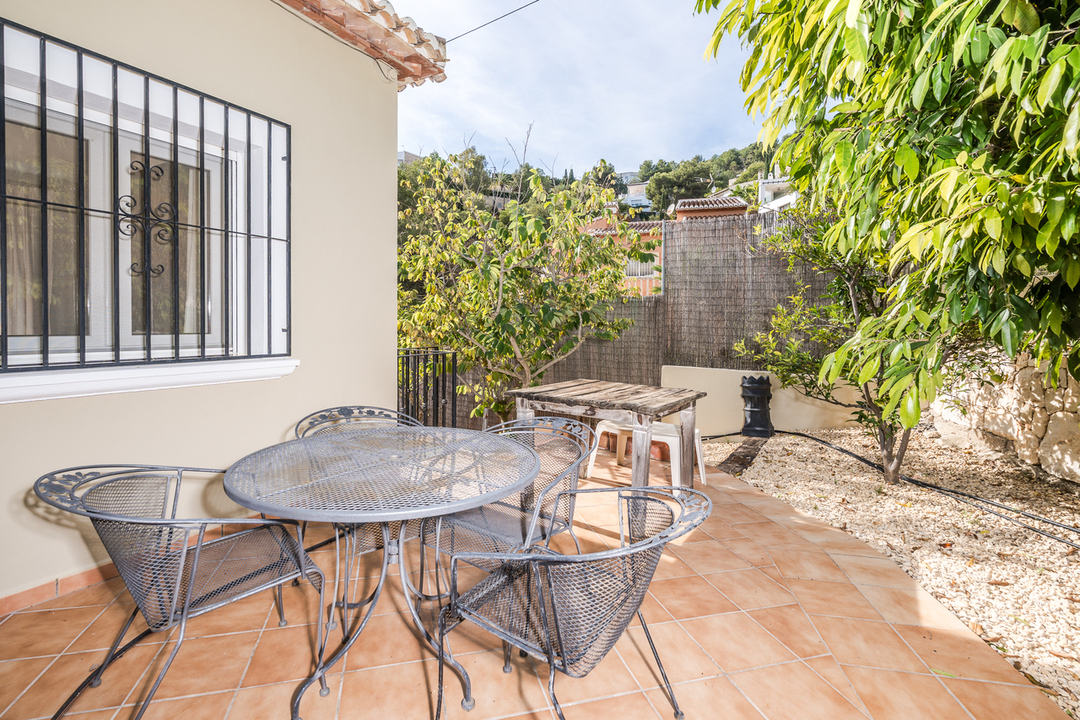 Terraza exterior con suelo de baldosas de terracota, conjunto de comedor de hierro forjado y ventana con rejas decorativas, rodeada de vegetación mediterránea y gravilla.