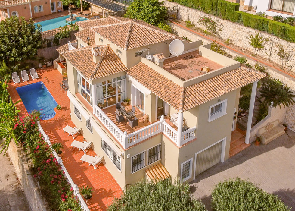 Vista aérea de una villa mediterránea con tejados de terracota, piscina privada y terrazas con balaustradas blancas y mobiliario de comedor exterior.