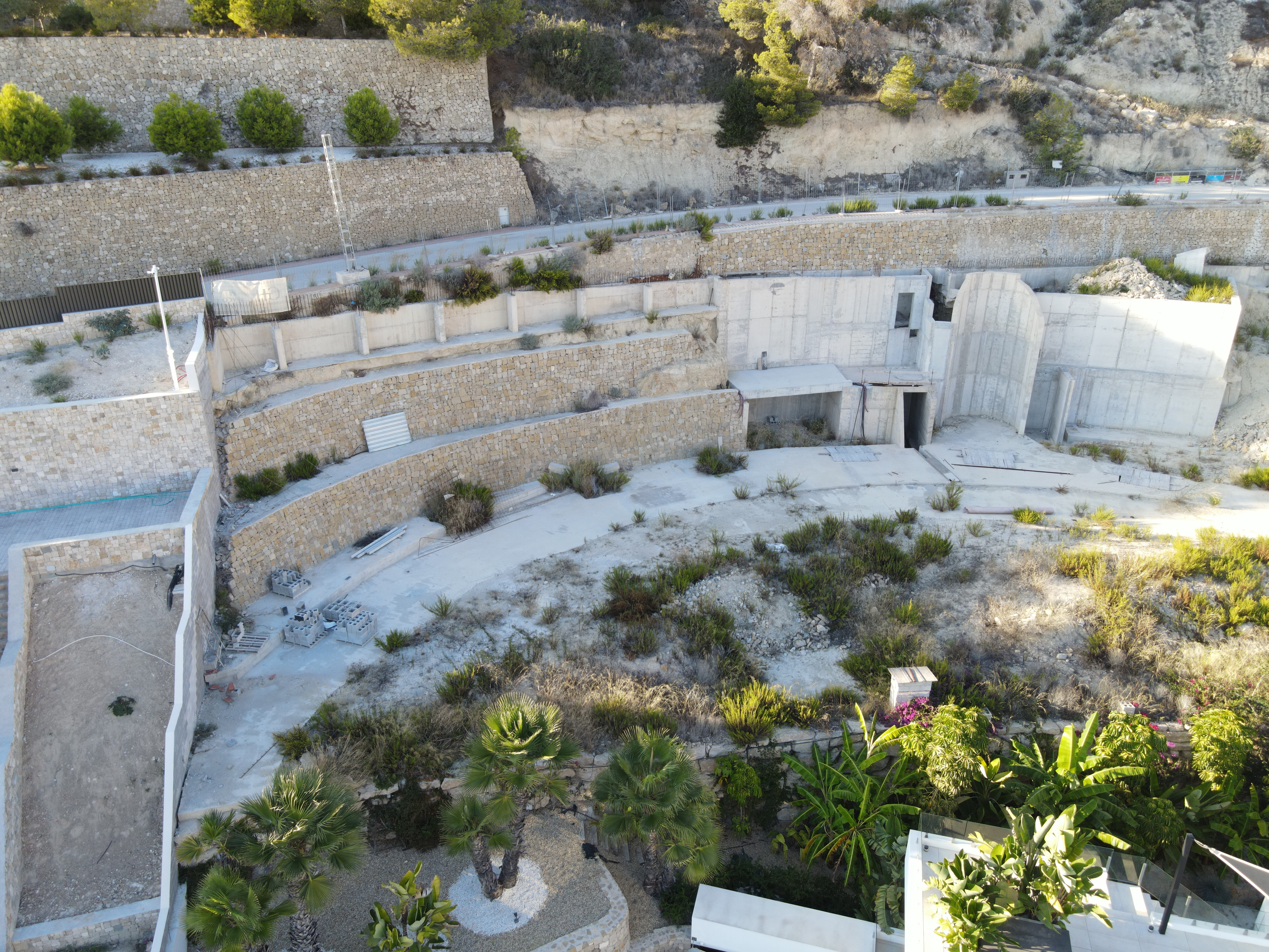 Vista aérea de muros de contención de piedra escalonados y estructuras de hormigón integradas en una ladera con palmeras y vegetación autóctona.