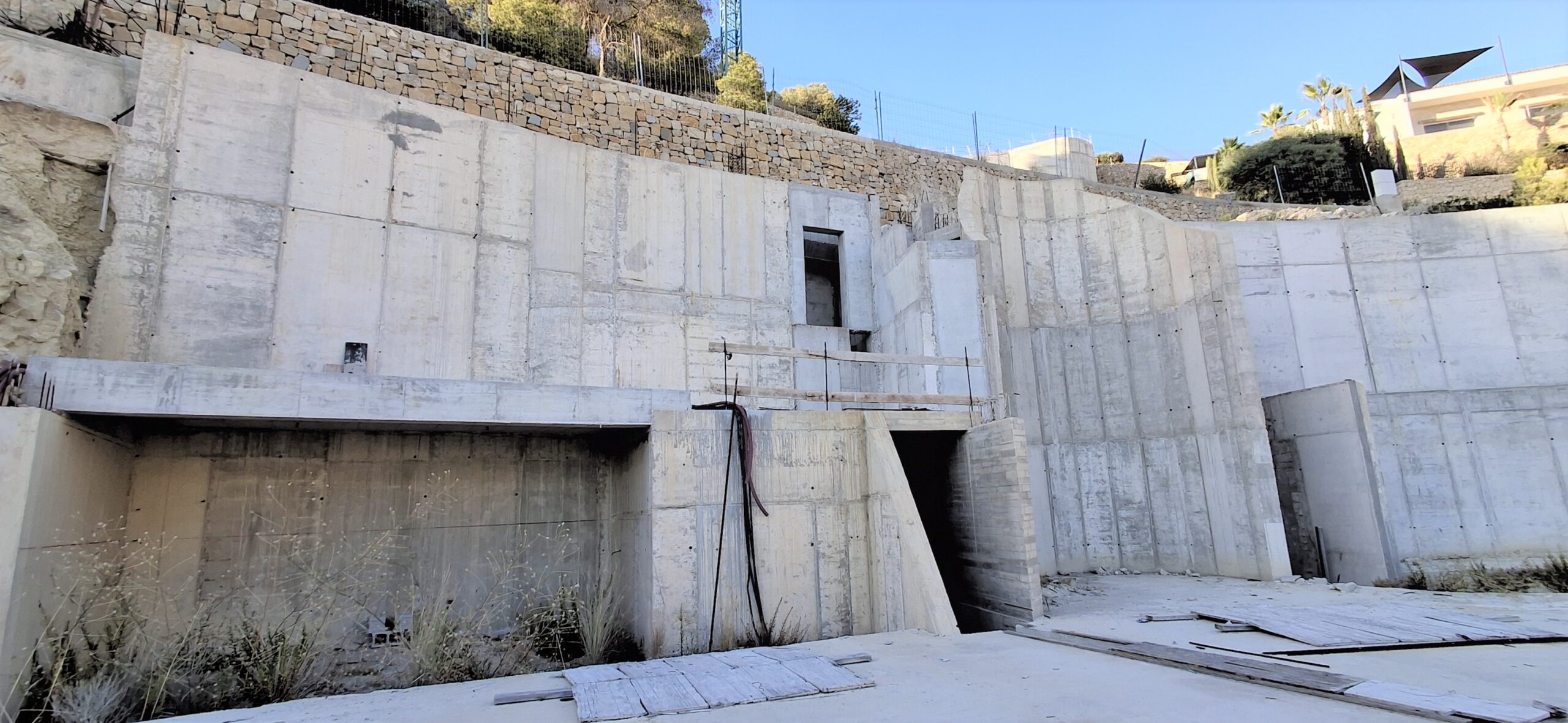 Vista estructural de los muros de contención de hormigón armado, con mampostería de piedra integrada en el nivel superior y huecos arquitectónicos en un terreno inclinado.