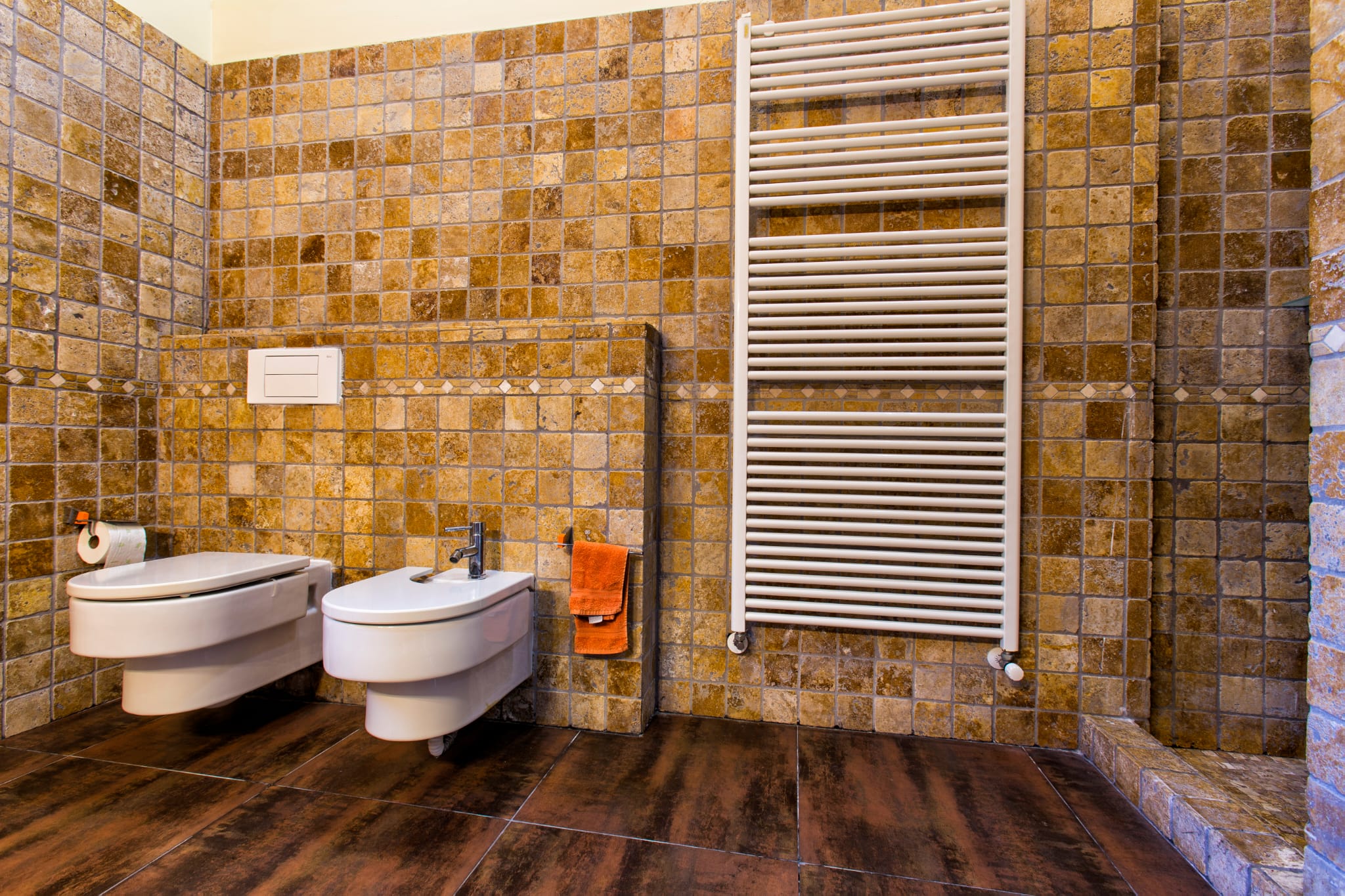 Baño con inodoro y bidé suspendidos, paredes revestidas en mosaico de piedra natural en tonos tierra y un radiador toallero blanco vertical.