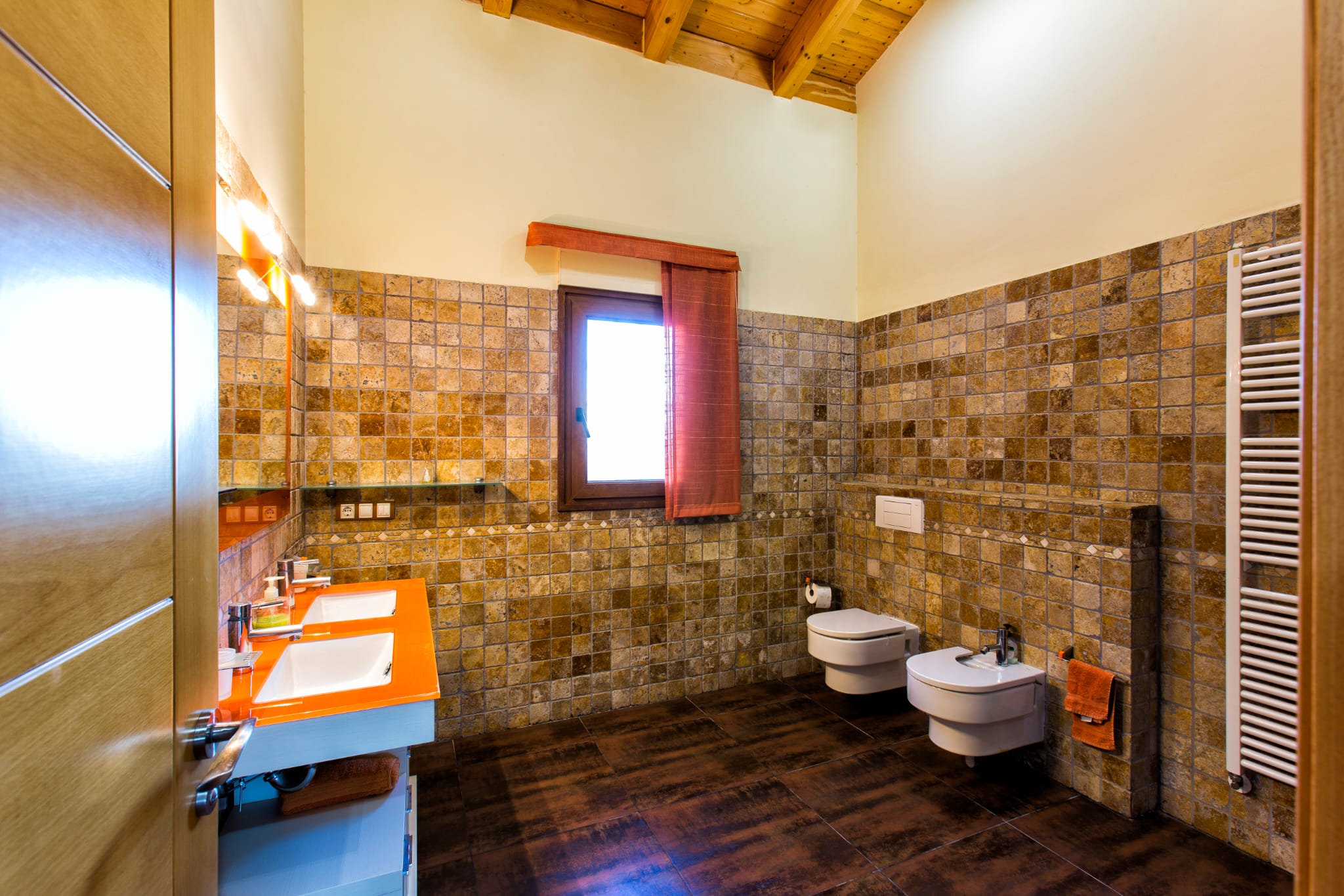Baño moderno con lavabo doble de encimera naranja, paredes revestidas en piedra natural y techo abovedado de madera con vigas vistas.