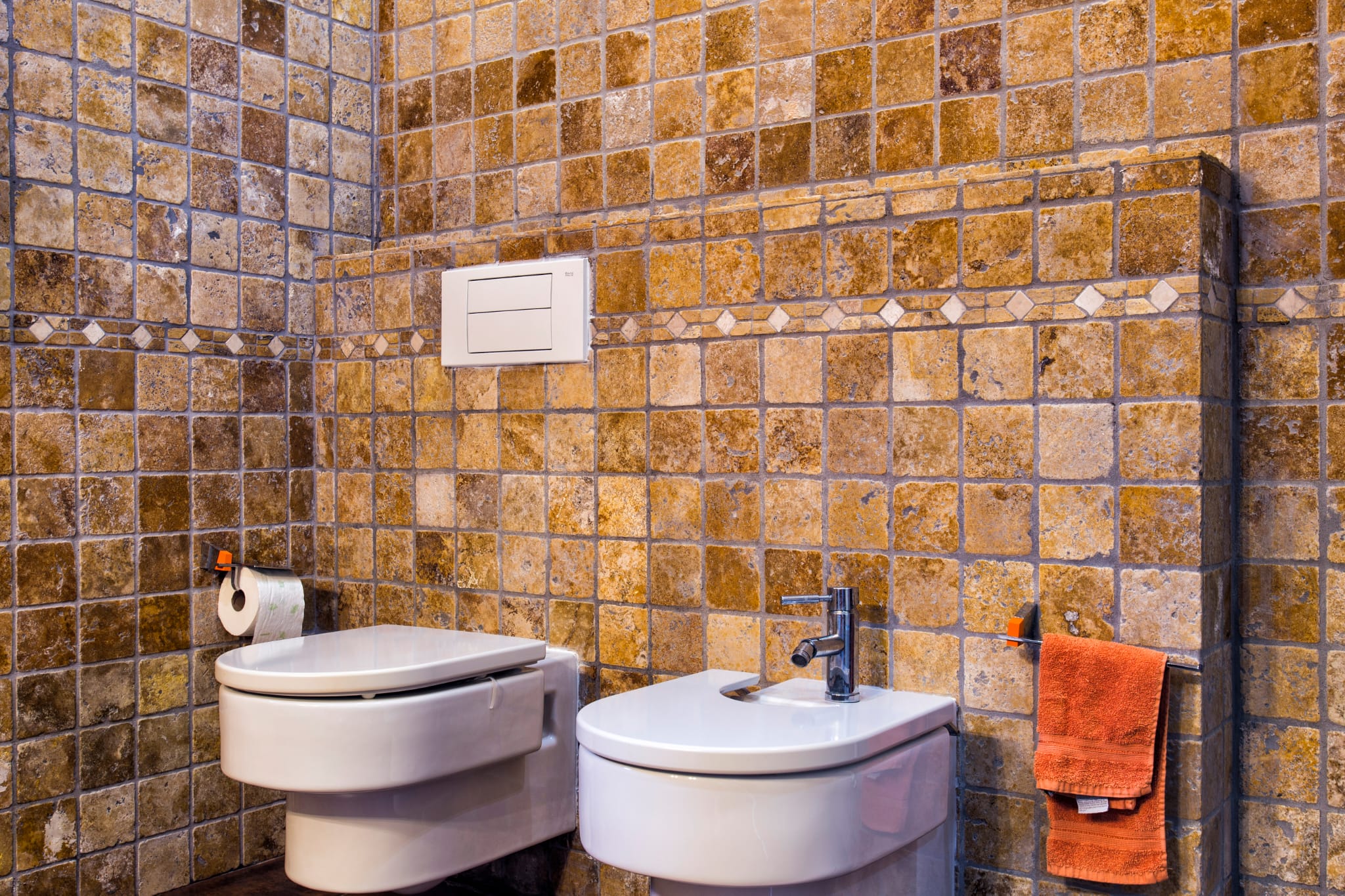 Baño revestido con azulejos rústicos en tonos ocre, inodoro y bidé suspendidos de cerámica blanca y grifería cromada.