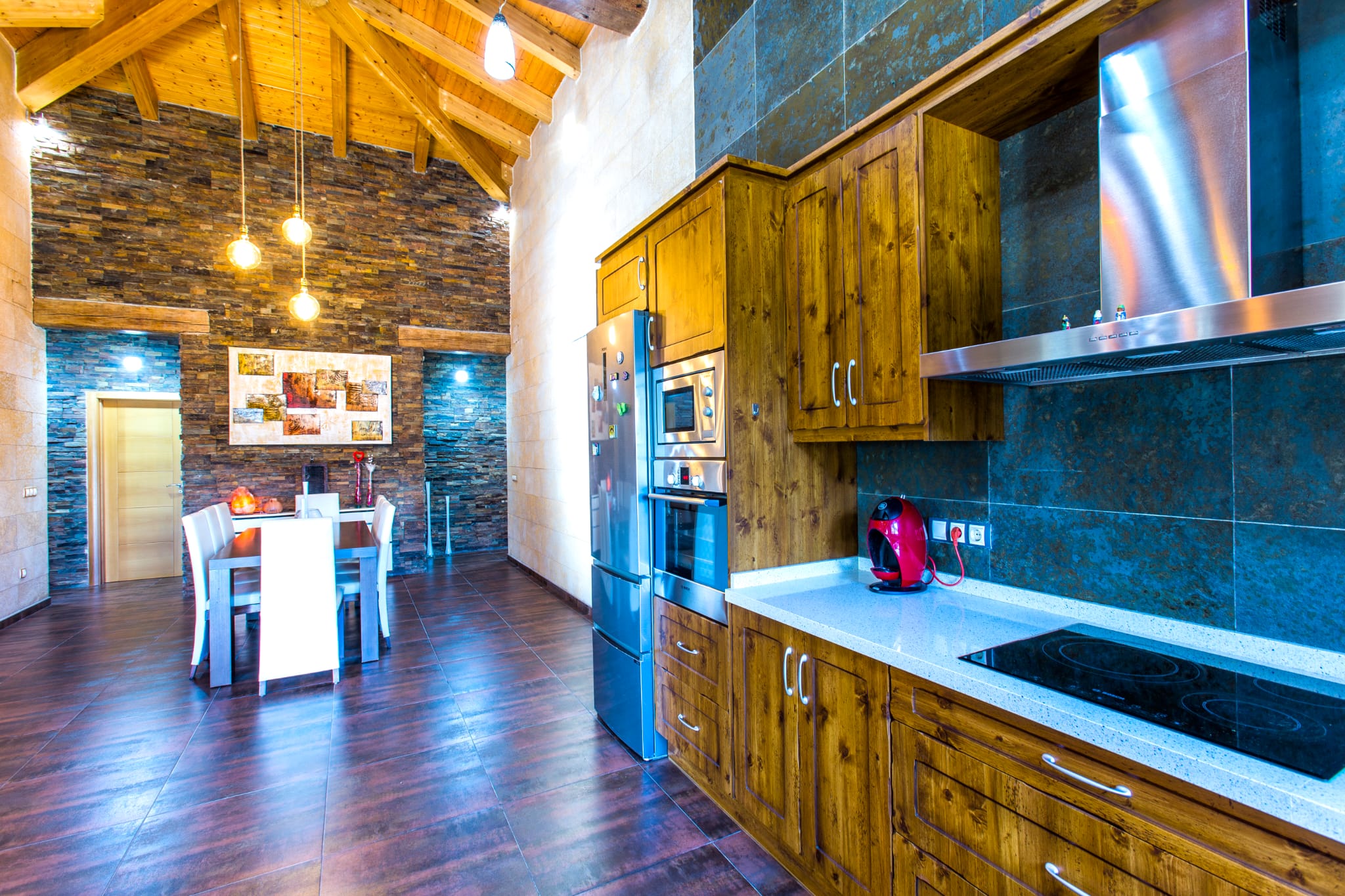 Cocina de estilo rústico con armarios de madera, electrodomésticos integrados y techo de vigas vistas. Destaca la pared de piedra natural que conecta con el comedor.