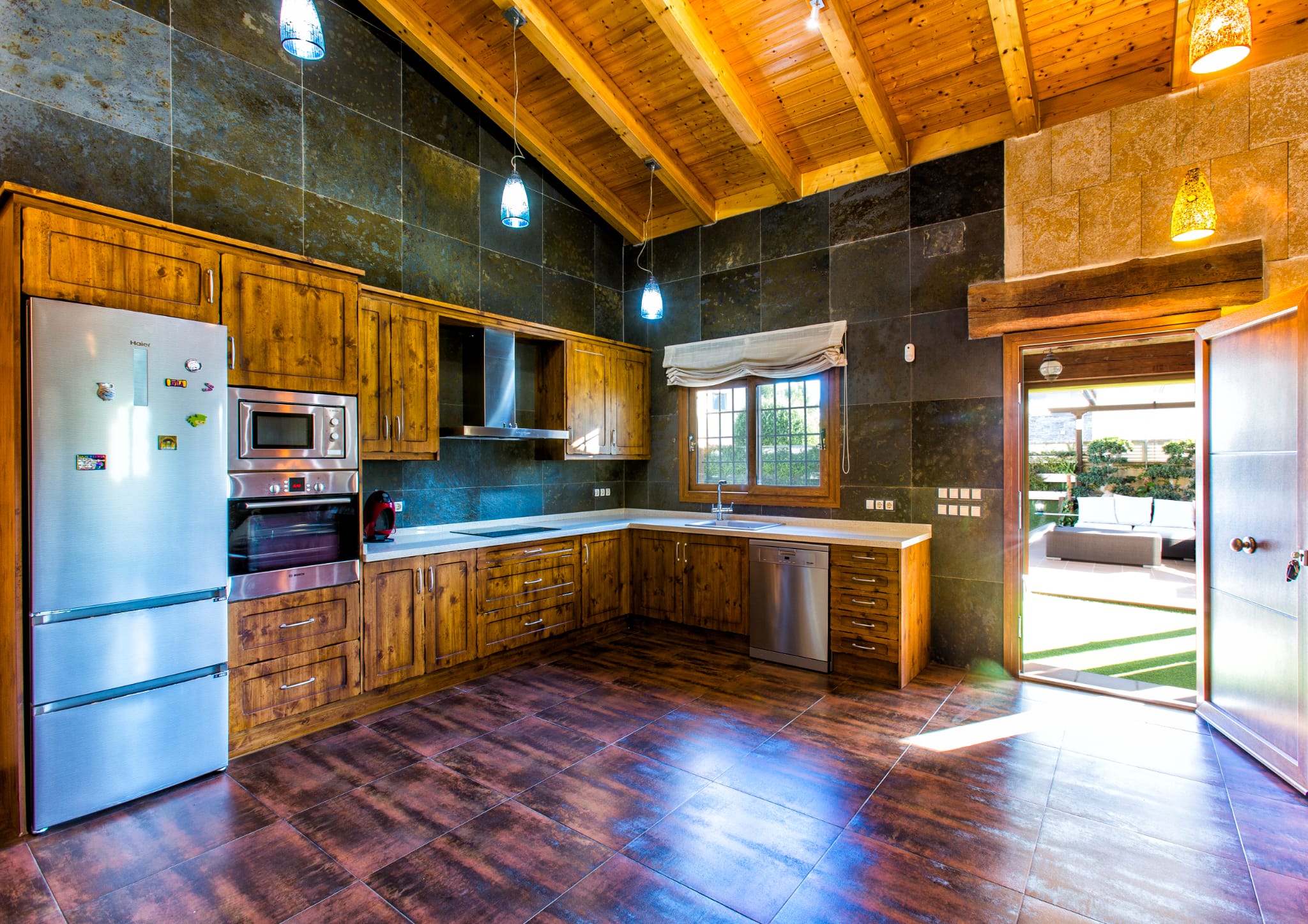 Cocina rústica con armarios de madera, paredes de pizarra oscura y techos de vigas vistas. Equipada con electrodomésticos de acero inoxidable y acceso directo al patio exterior.