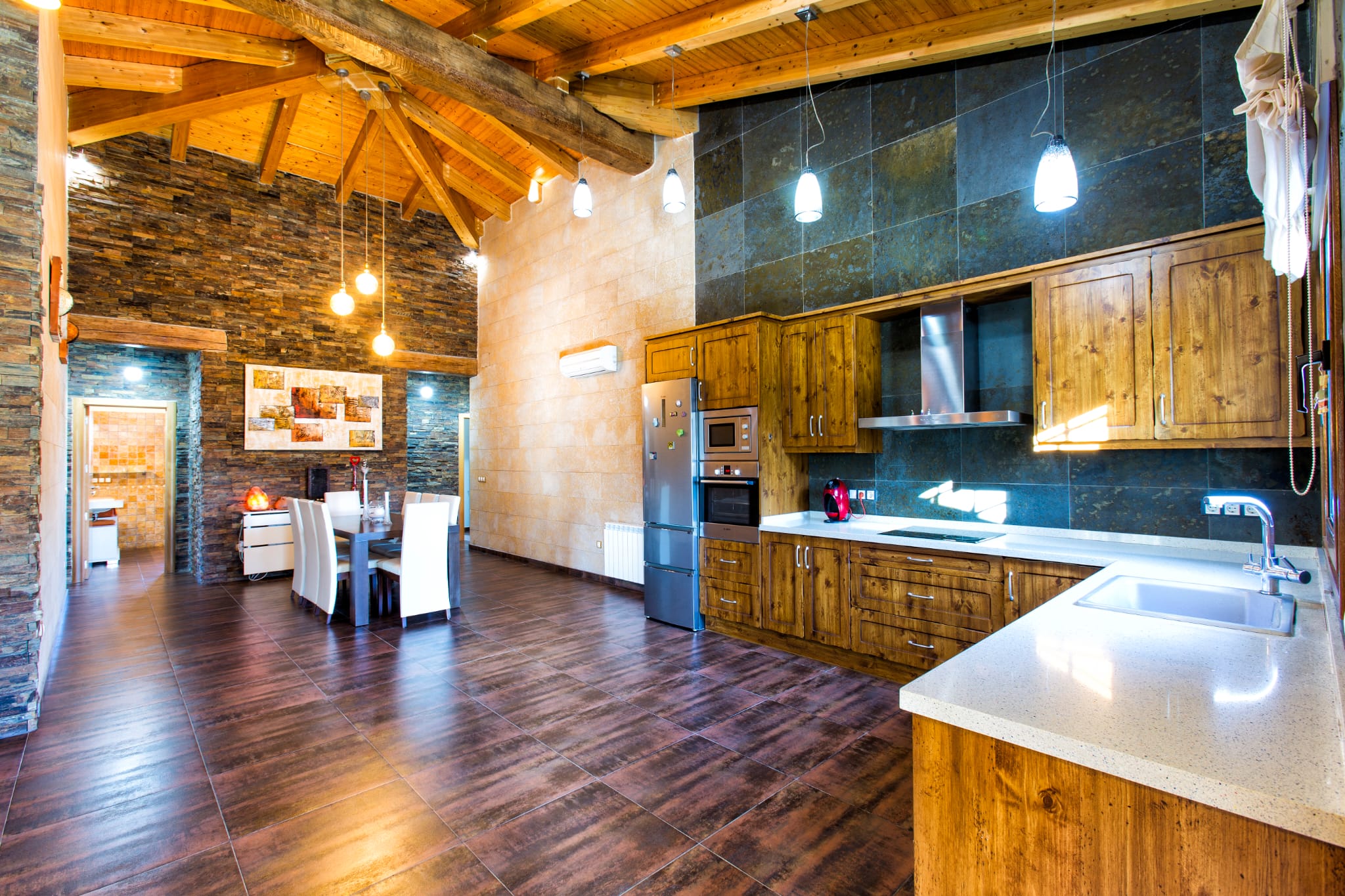 Cocina y comedor de planta abierta con techos altos de madera, paredes de piedra natural, mobiliario rústico y electrodomésticos de acero inoxidable.