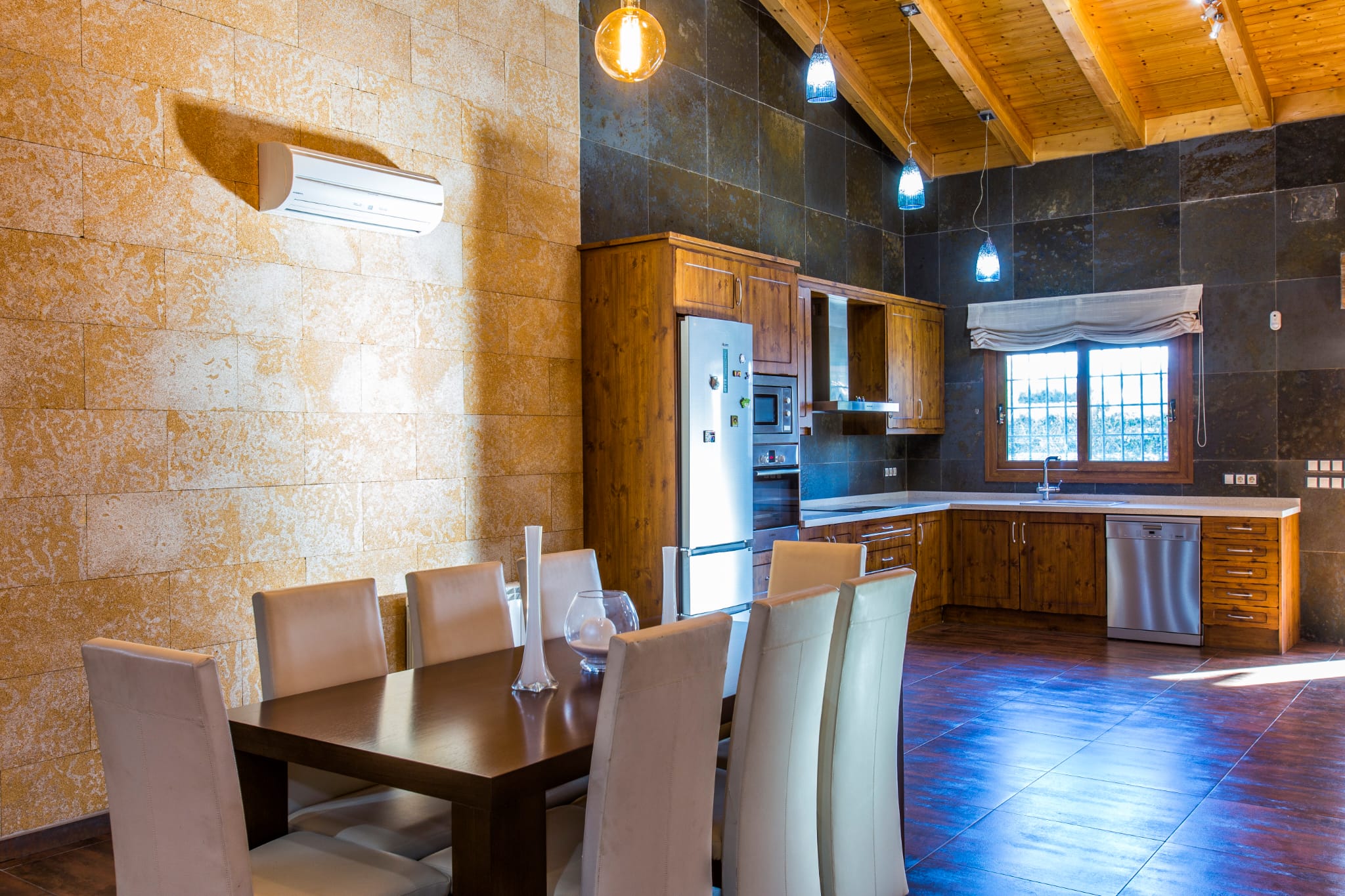Cocina y comedor de planta abierta con techos altos de madera, paredes de piedra natural, mobiliario rústico y electrodomésticos integrados de acero inoxidable.