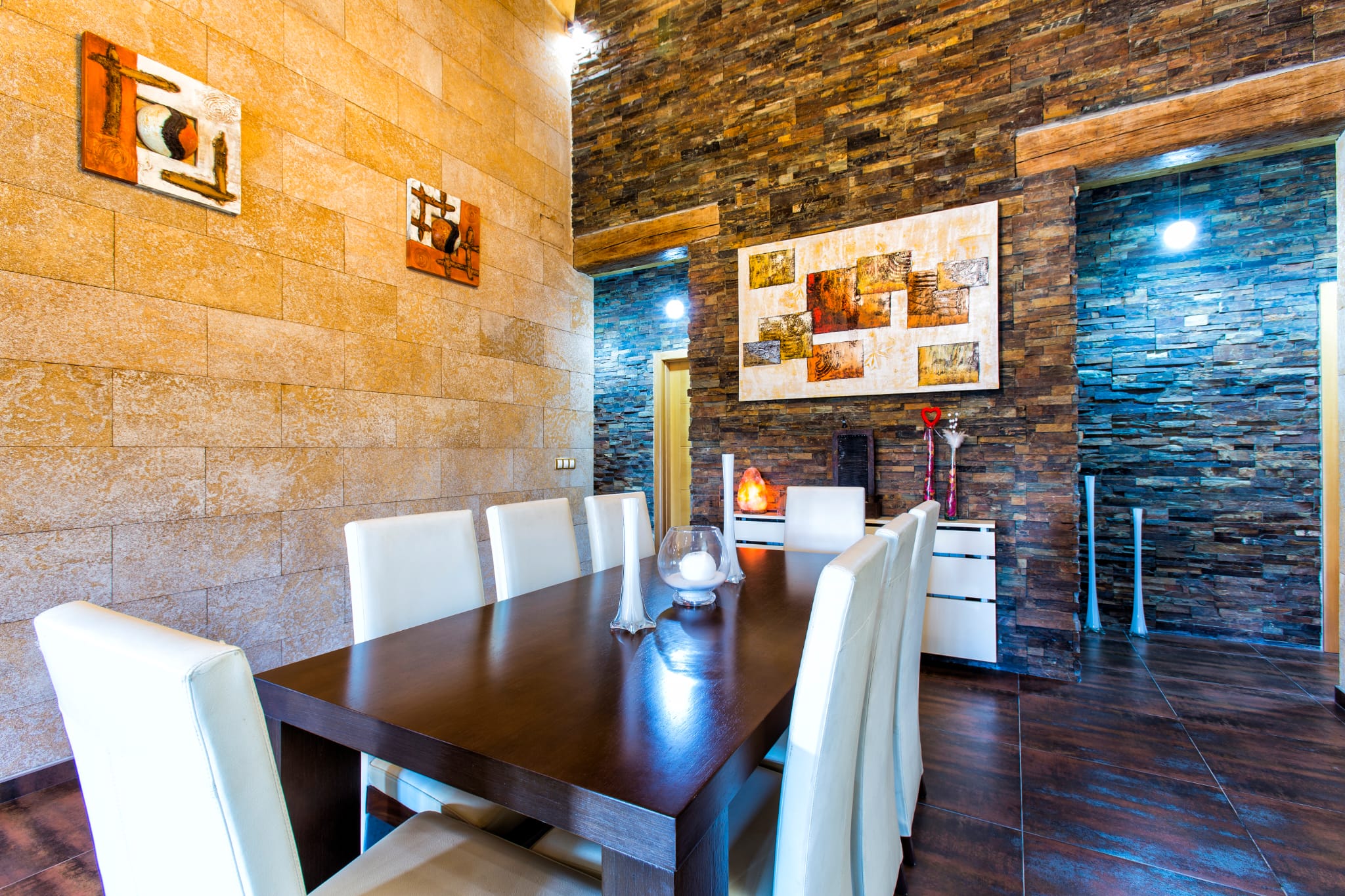 Comedor con mesa de madera oscura y sillas blancas, destacando paredes de piedra natural y pizarra con obras de arte abstractas.