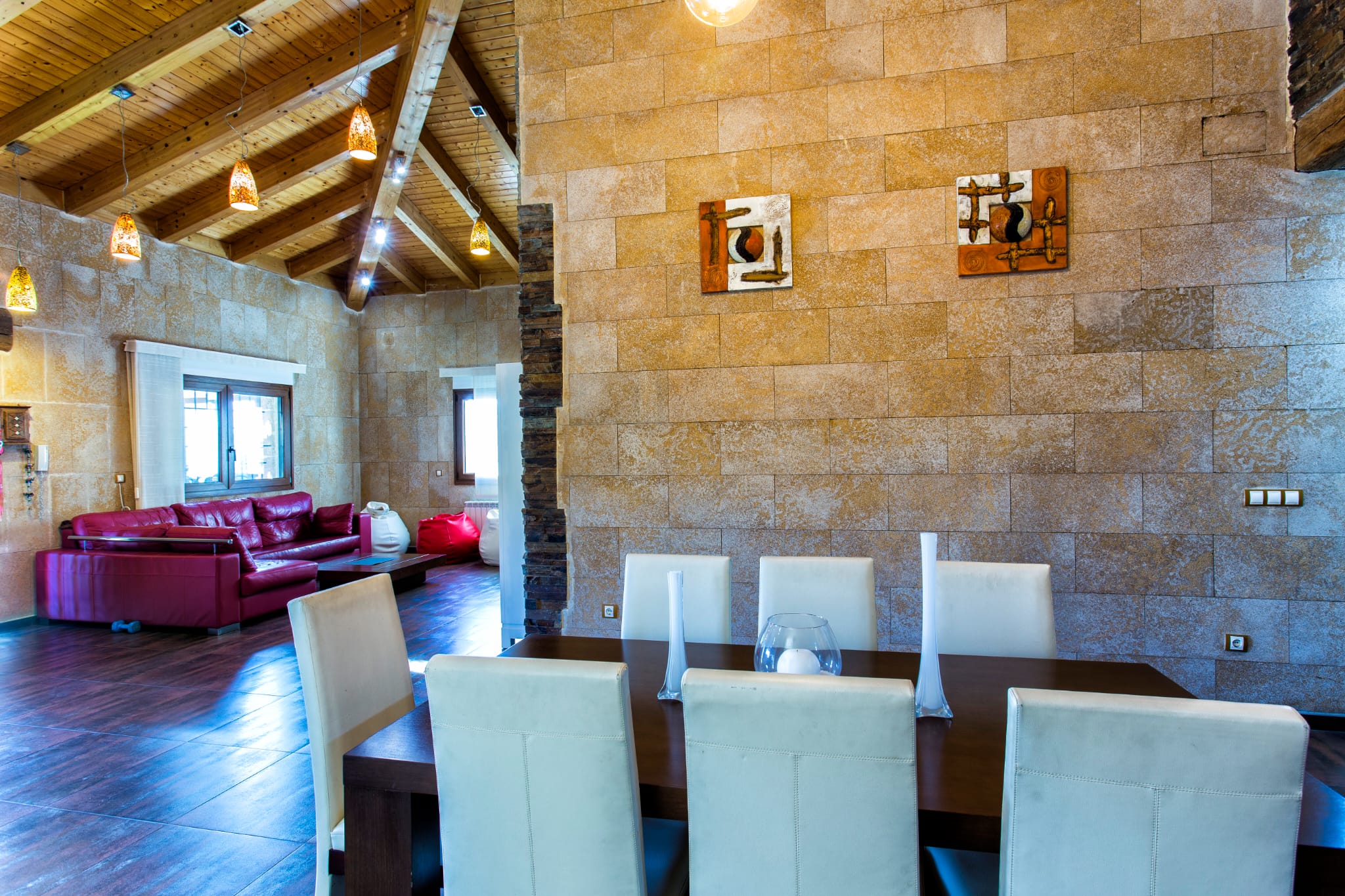 Comedor de planta abierta con paredes de piedra natural, techos altos de madera con vigas vistas y mobiliario moderno integrado con la zona de estar.