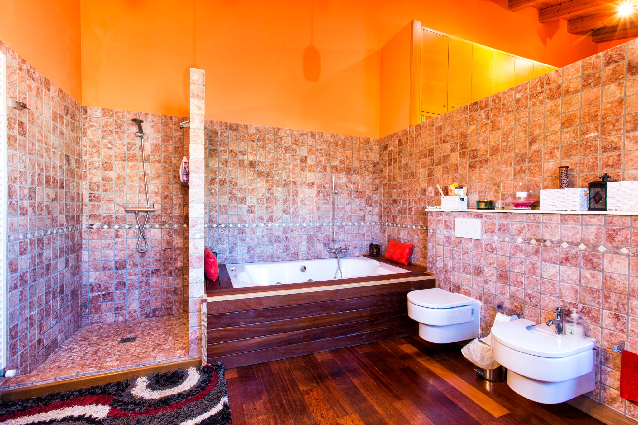 Cuarto de baño con ducha de obra en piedra, bañera revestida en madera oscura y sanitarios suspendidos. Destacan las paredes en tono naranja y el suelo de madera noble.