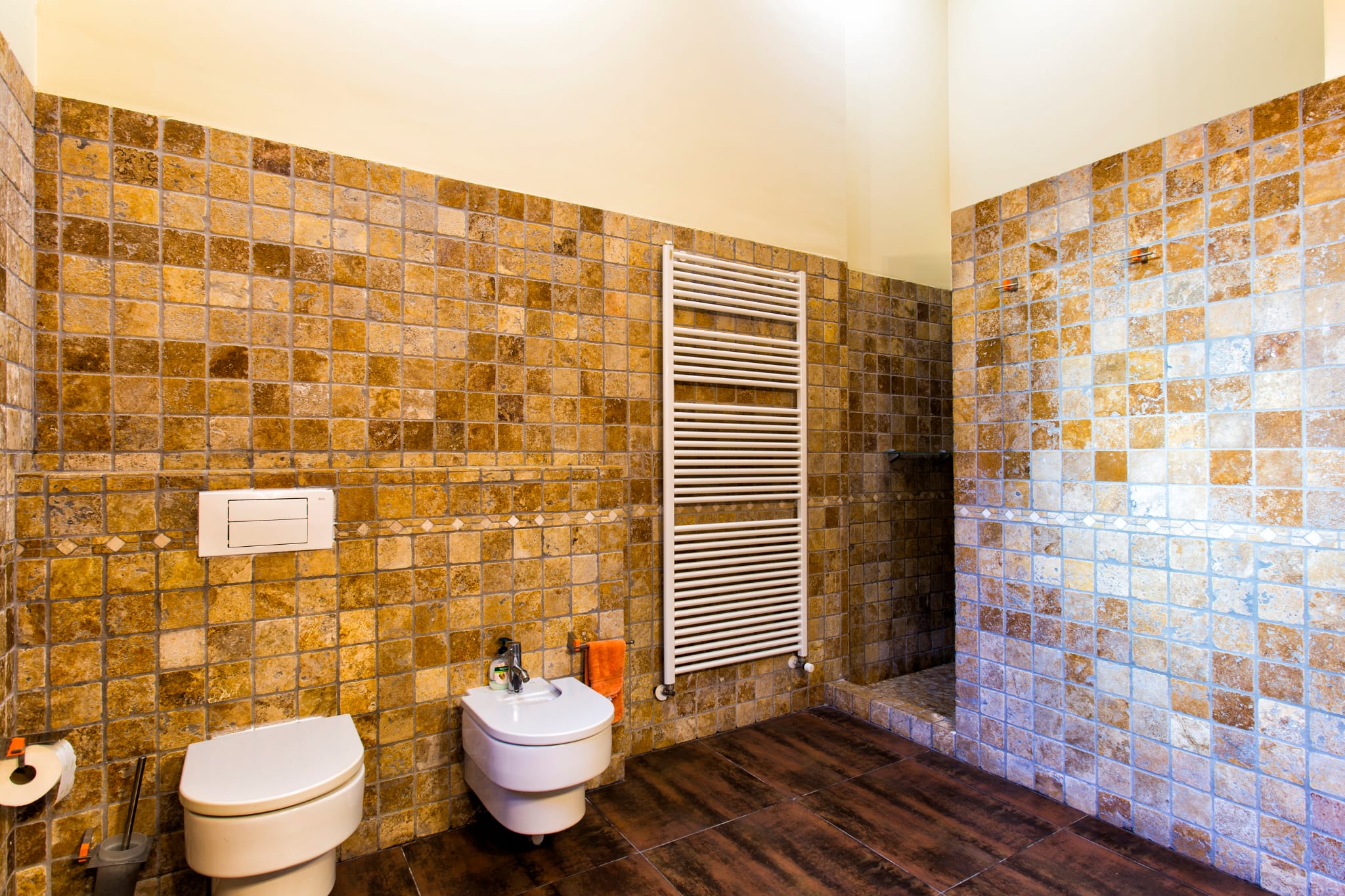 Cuarto de baño con paredes de mosaico en tonos tierra, sanitarios suspendidos, radiador toallero blanco y suelo cerámico de efecto madera oscura.