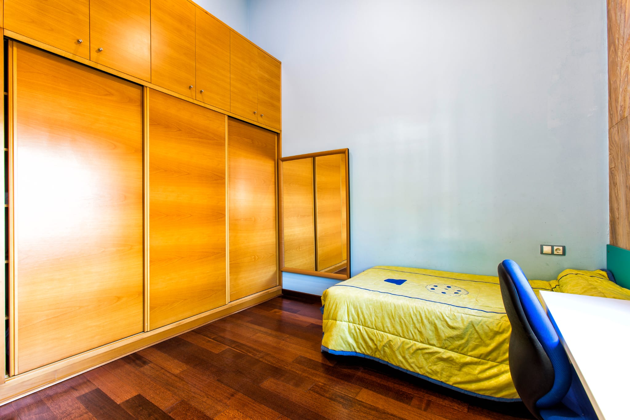 Dormitorio con amplio armario empotrado de madera, puertas correderas, suelo de parqué oscuro y cama individual junto a una zona de escritorio.