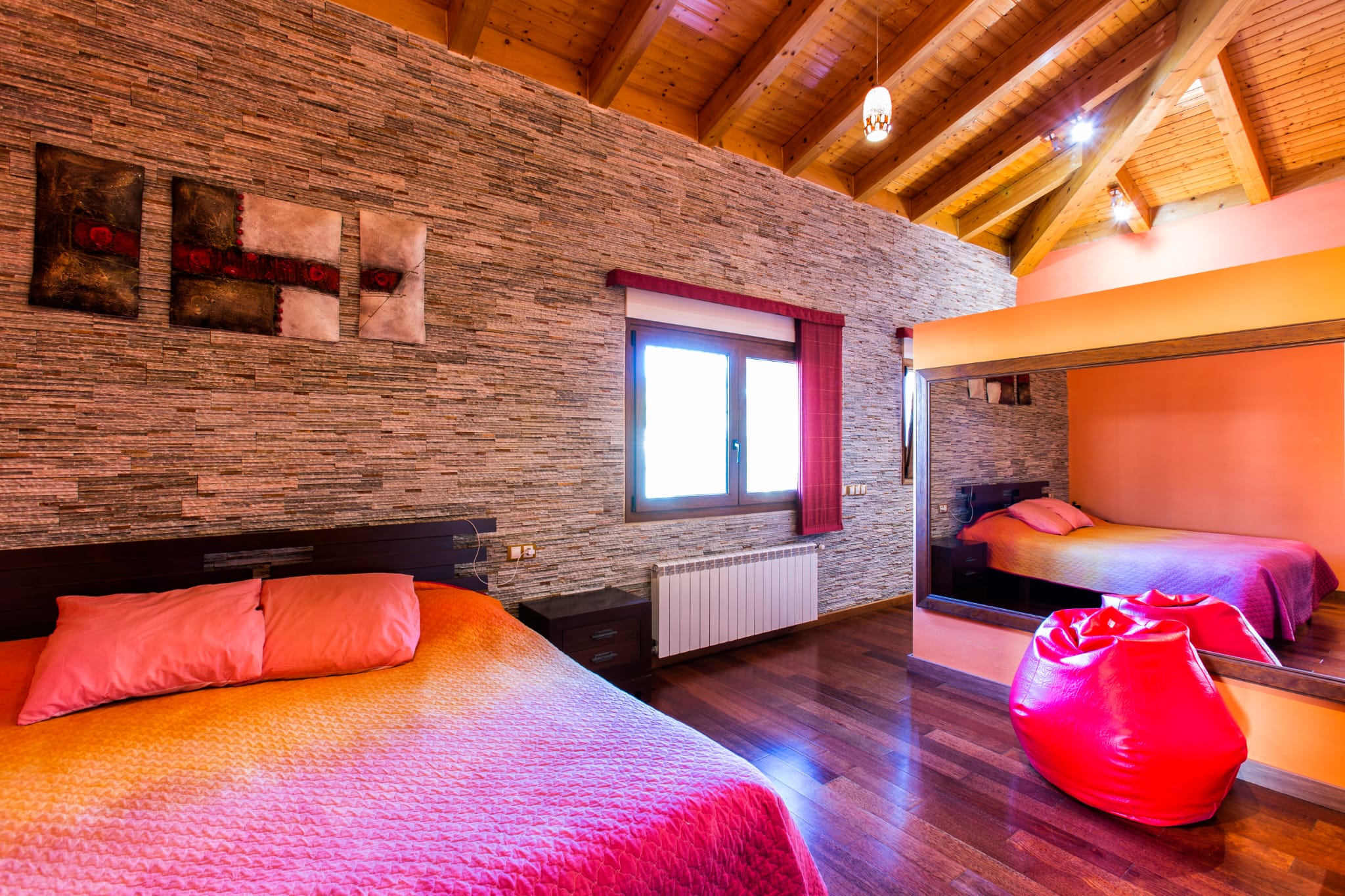 Dormitorio con pared de piedra texturizada, techos abovedados de vigas de madera y suelos oscuros. Un gran espejo refleja la cama con textiles en tonos fucsia y naranja.