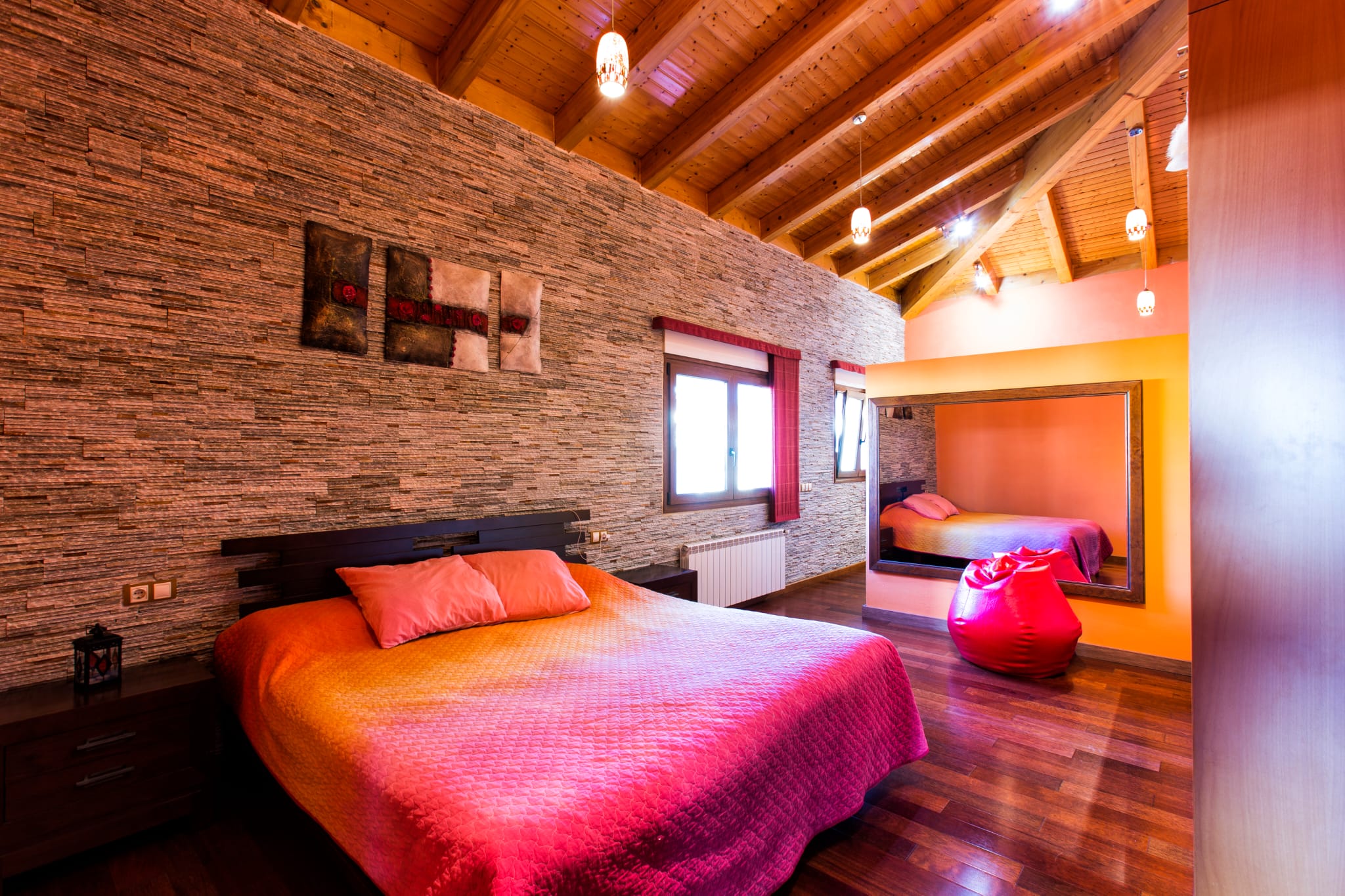 Dormitorio con pared de piedra texturizada, techos de vigas de madera inclinadas y suelo de parqué oscuro. Destaca un gran espejo mural y lámparas colgantes.