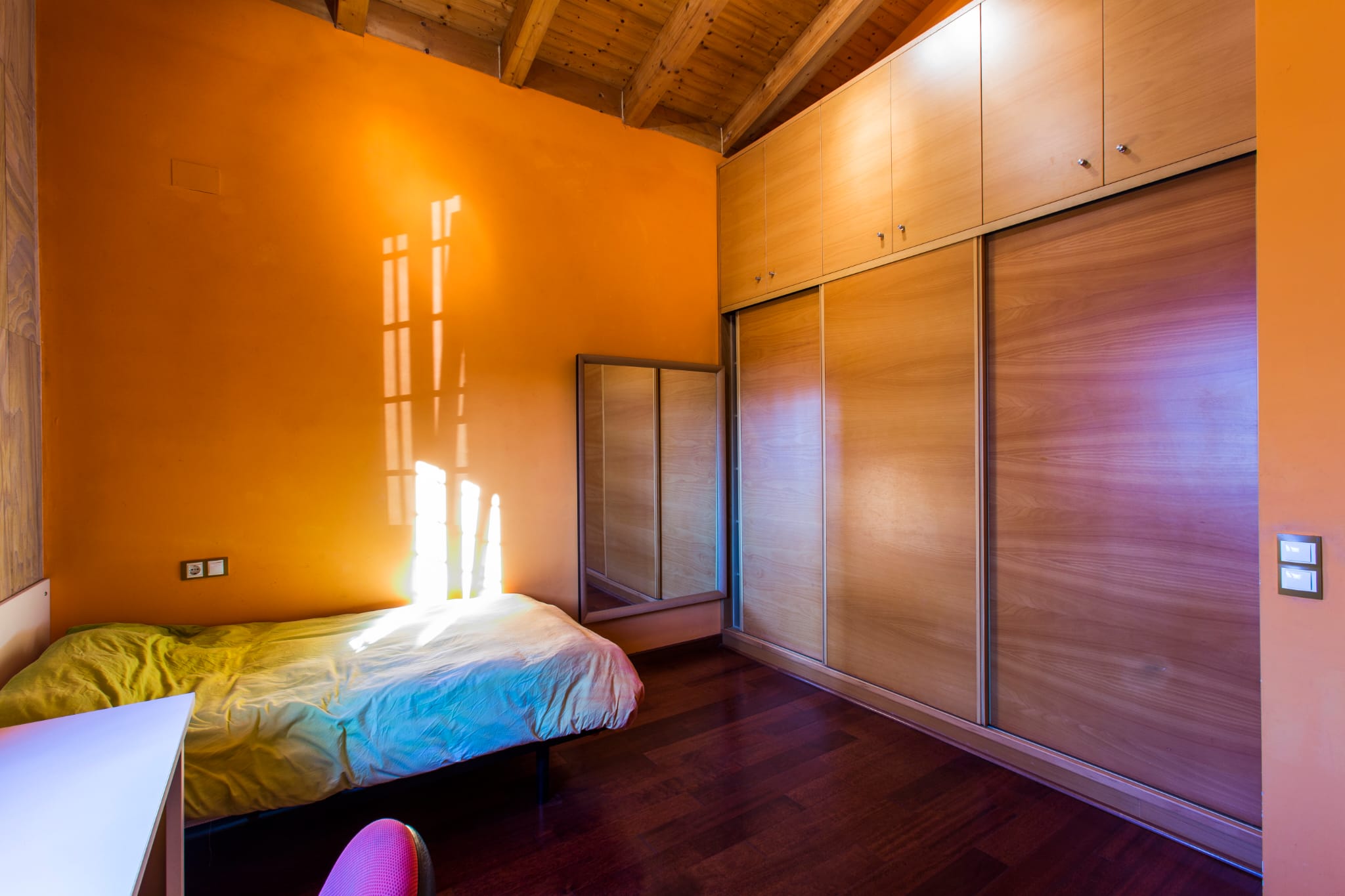 Dormitorio con paredes naranjas, techos de madera con vigas vistas y un amplio armario empotrado de madera con puertas correderas.