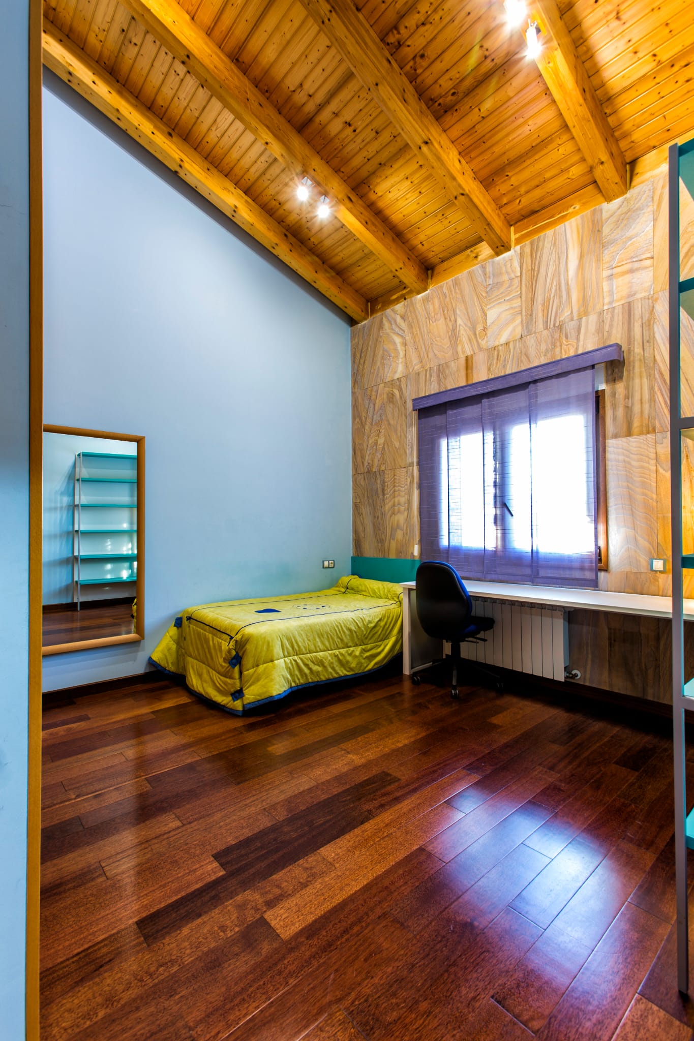 Dormitorio con techos de vigas de madera, suelos de parqué oscuro, zona de estudio con escritorio blanco y una pared de paneles con textura natural.