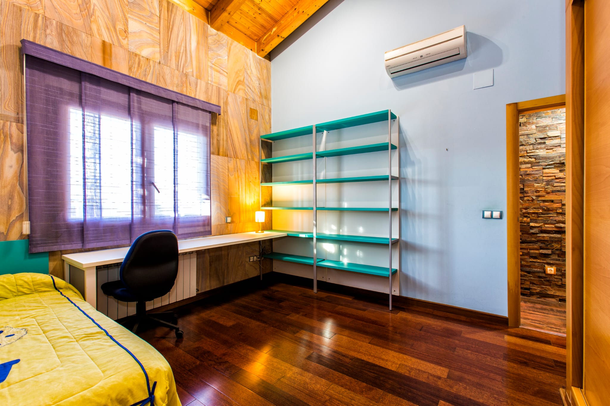Dormitorio con zona de estudio, techos de madera inclinados, estantería turquesa y escritorio bajo ventana con estores púrpuras.