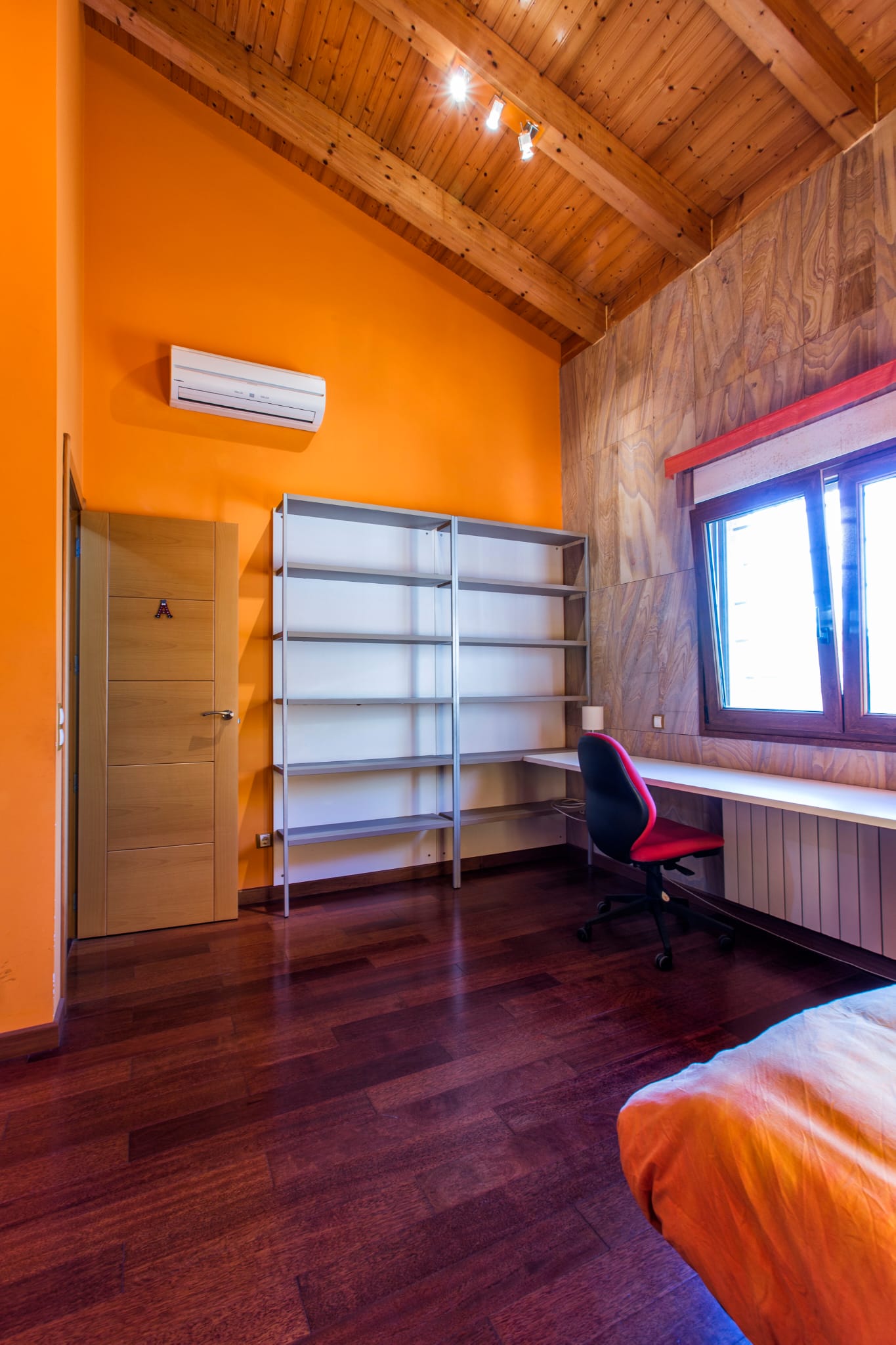 Habitación con techos altos de madera vista, paredes en tono naranja, zona de escritorio integrada, estantería metálica y suelos de madera oscura.