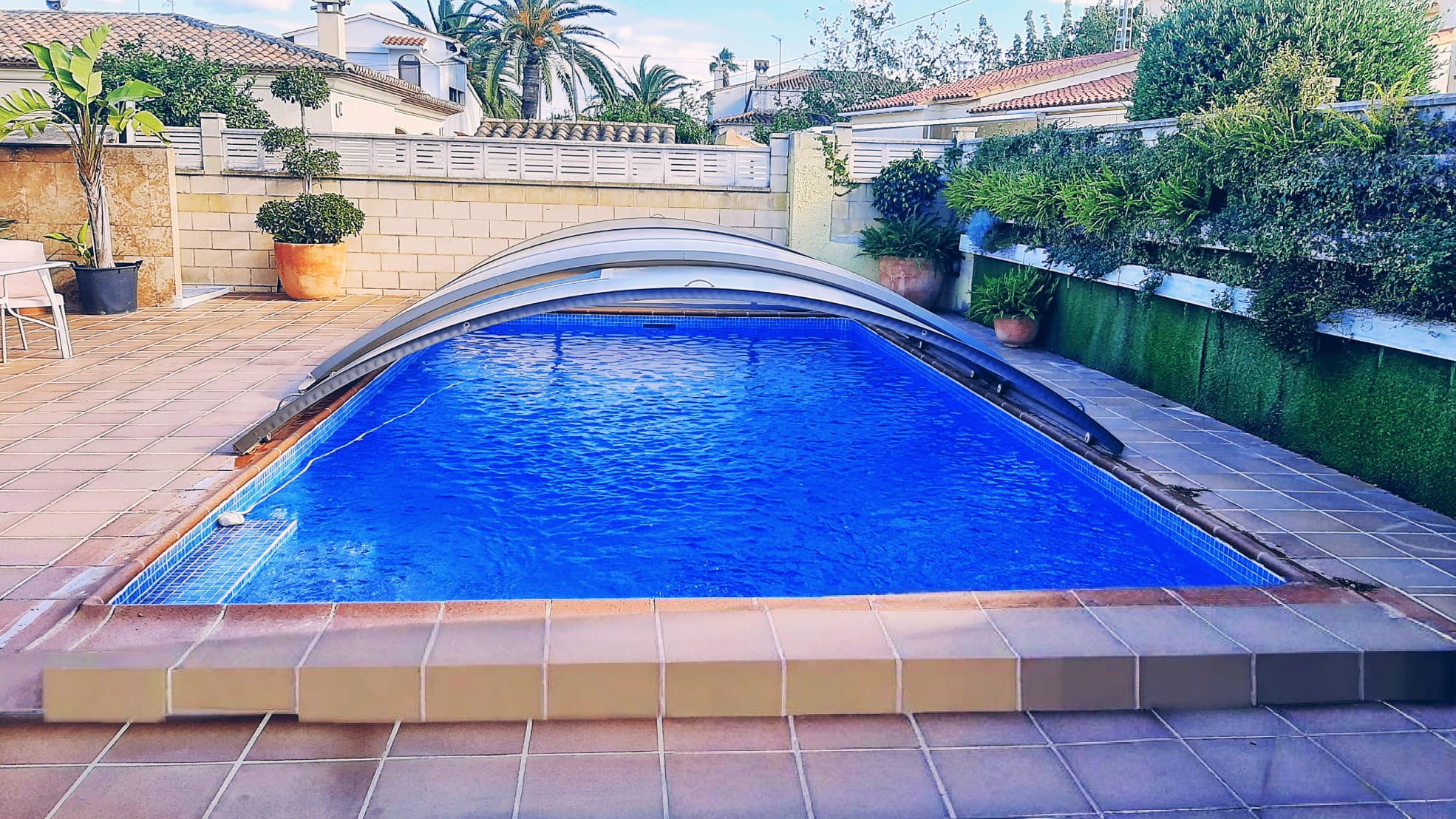 Piscina privada con cubierta telescópica y revestimiento de gresite azul, rodeada por una terraza de gres y muros perimetrales con vegetación ornamental.